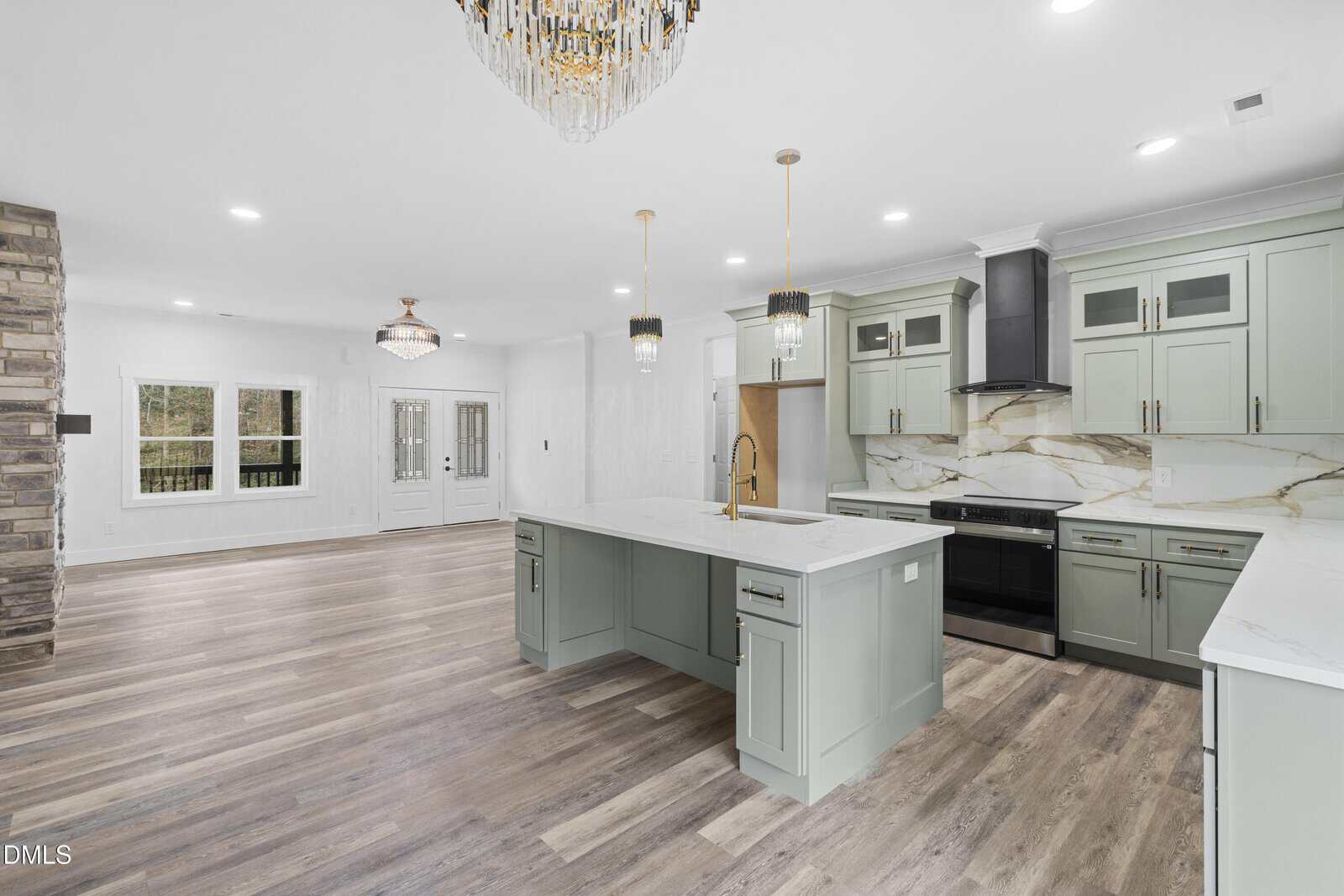 Modern kitchen with light green cabinets, marble countertops, gold fixtures, wood-style flooring, and open layout to living area with stone accent wall and large windows. Elegant chandeliers hang from the ceiling.