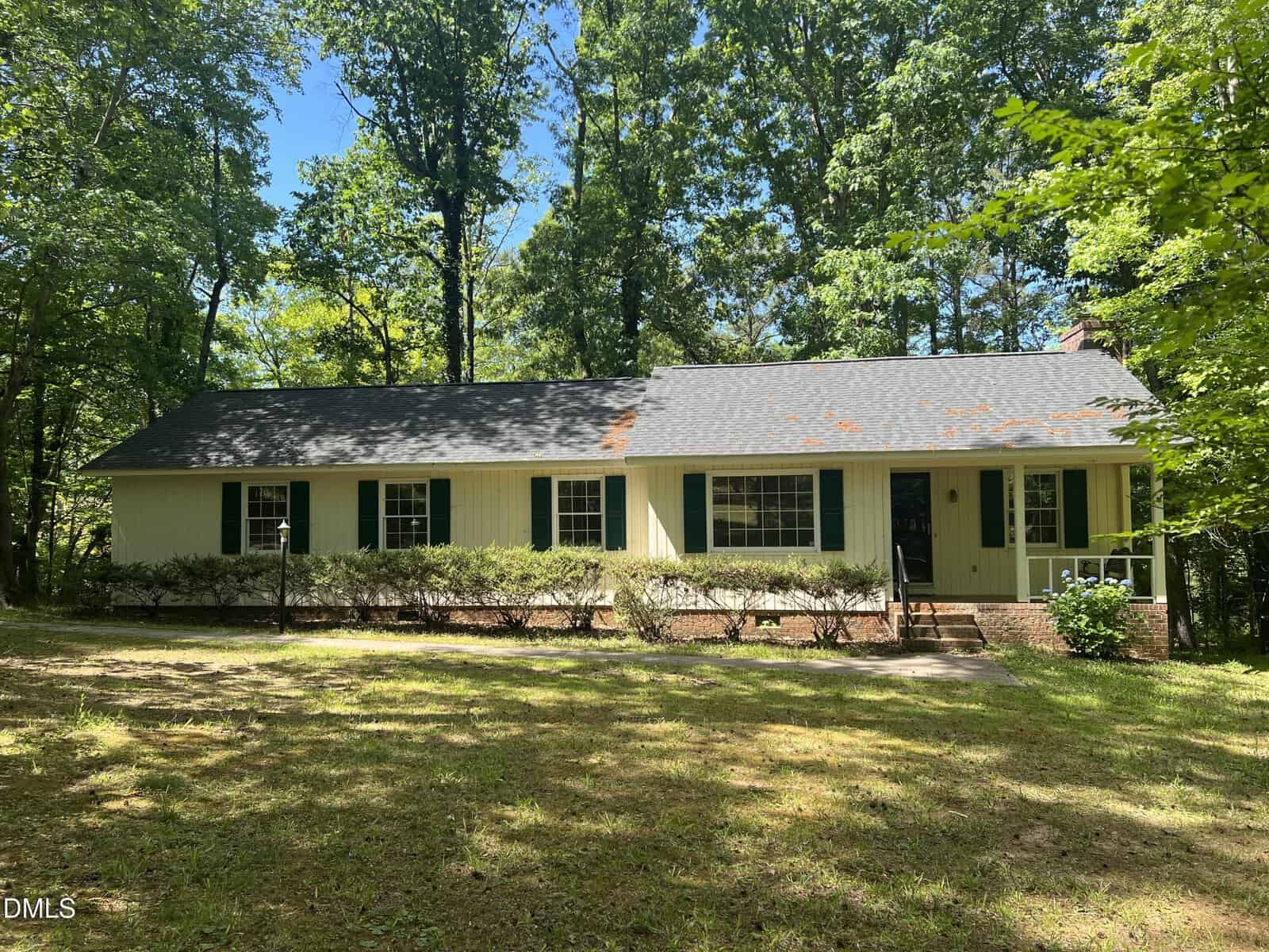 Single-story yellow house with dark green shutters and a front porch, surrounded by tall trees and shaded by their branches. There is a grassy yard in front and sunlight filtering through the leaves.