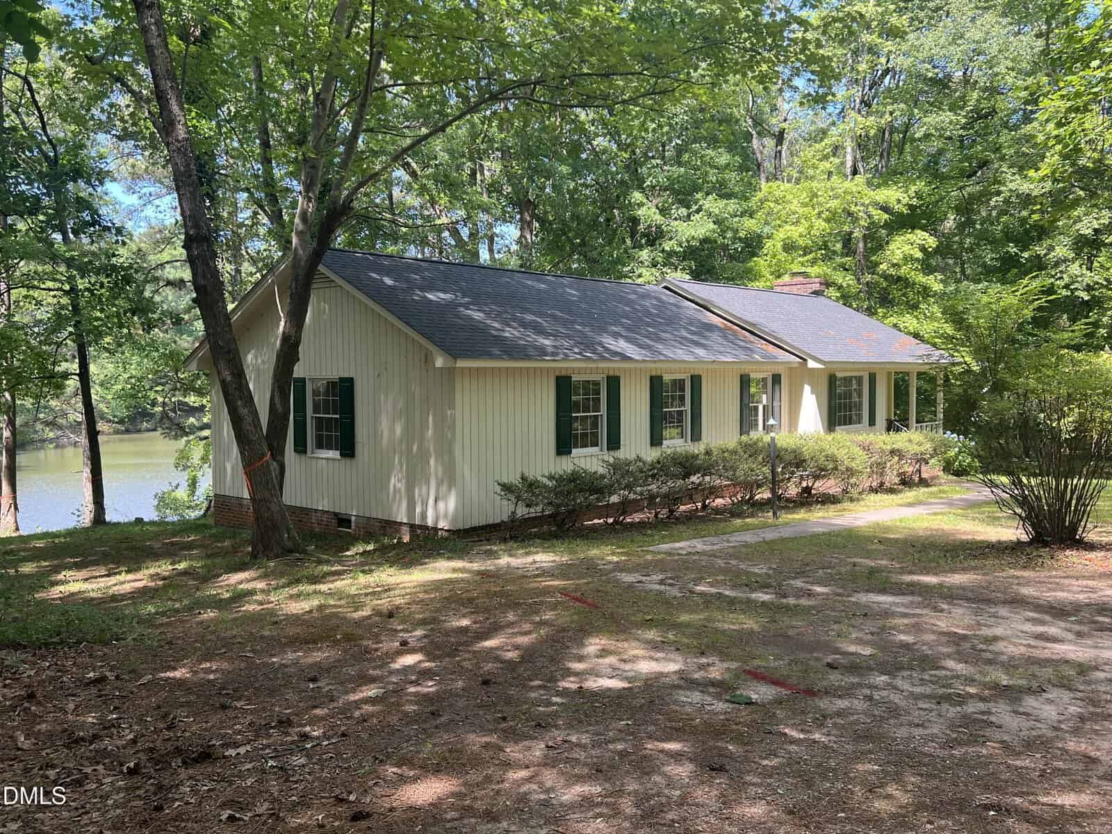 Nestled at 113 Country Brook Lane in Youngsville, this single-story house with light siding and green shutters sits beside a tranquil lake, surrounded by trees and bushes with sunlight filtering through the leaves.
