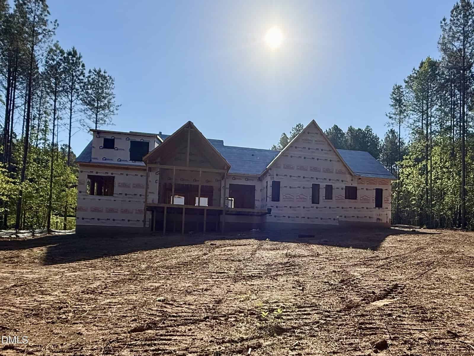 A house under construction, wrapped in protective sheeting, sits on a cleared dirt lot surrounded by tall pine trees with the sun shining brightly overhead.
