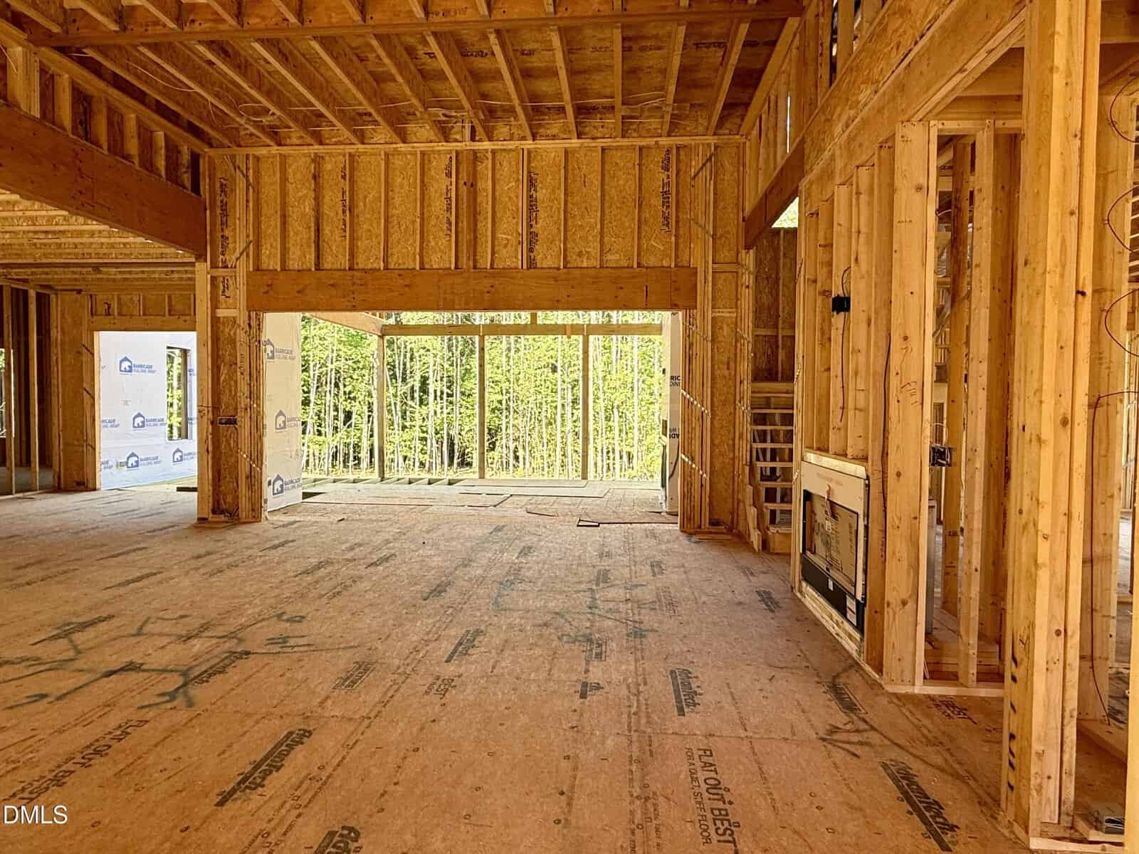 Interior view of a house under construction, showing exposed wooden framing, unfinished walls, large windows, and open floor plan with ample natural light.