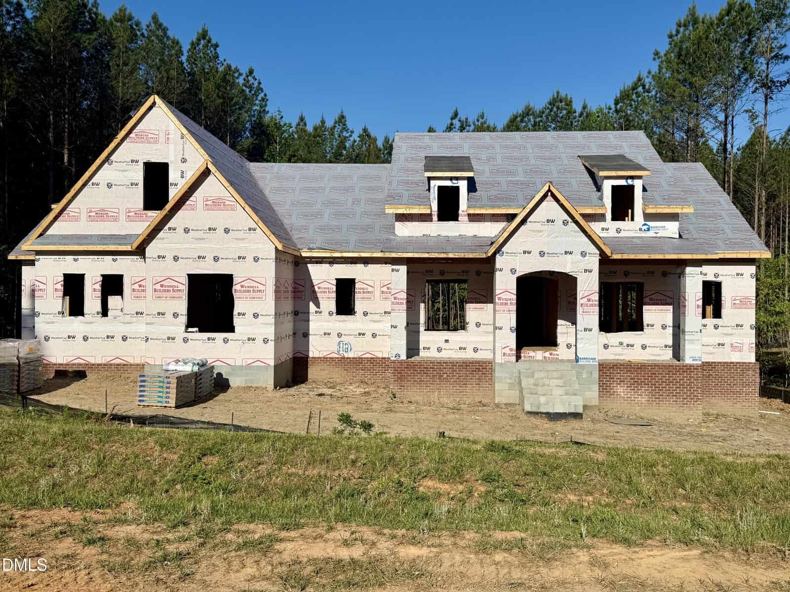 A house under construction with exposed framing, roof sheathing, and house wrap visible. The structure has two dormer windows and a brick foundation, surrounded by dirt and a grassy area, with trees in the background.