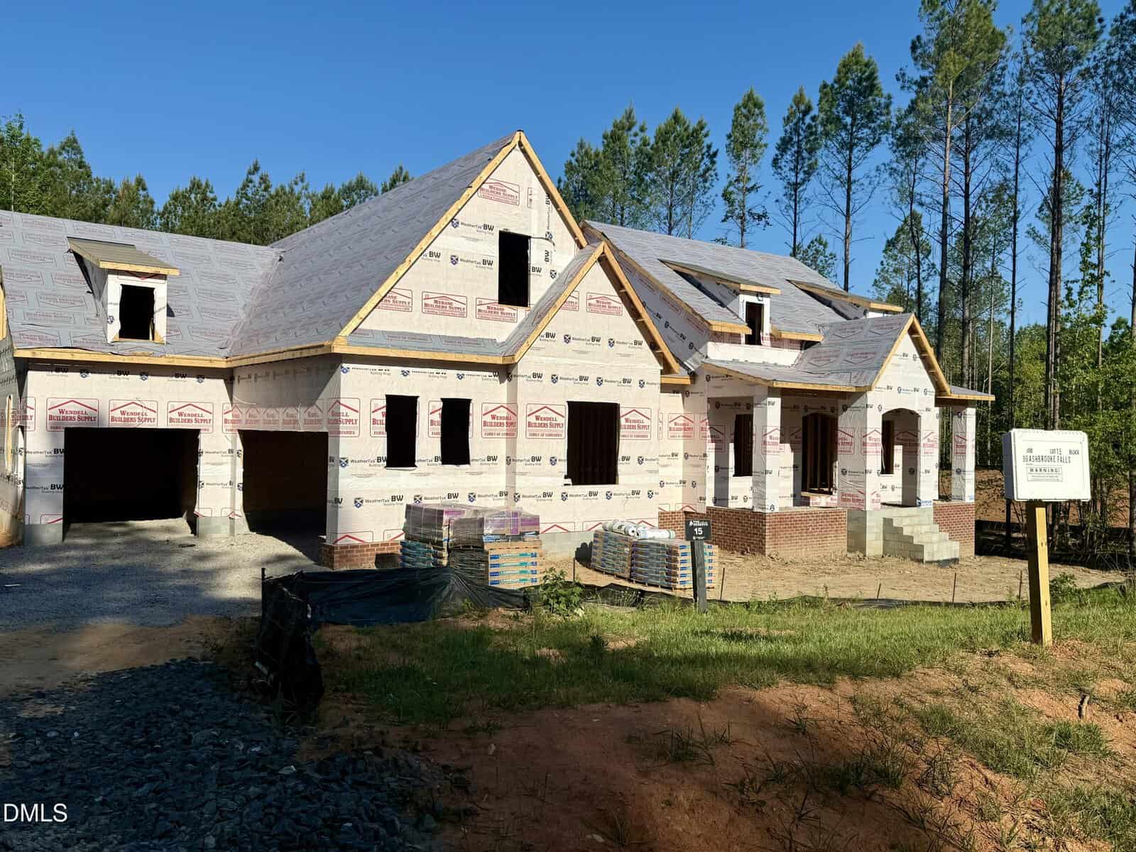 A large house under construction with exposed plywood, house wrap, unfinished roof, and brick steps. Construction materials and pallets are on the ground, surrounded by trees and a clear blue sky.