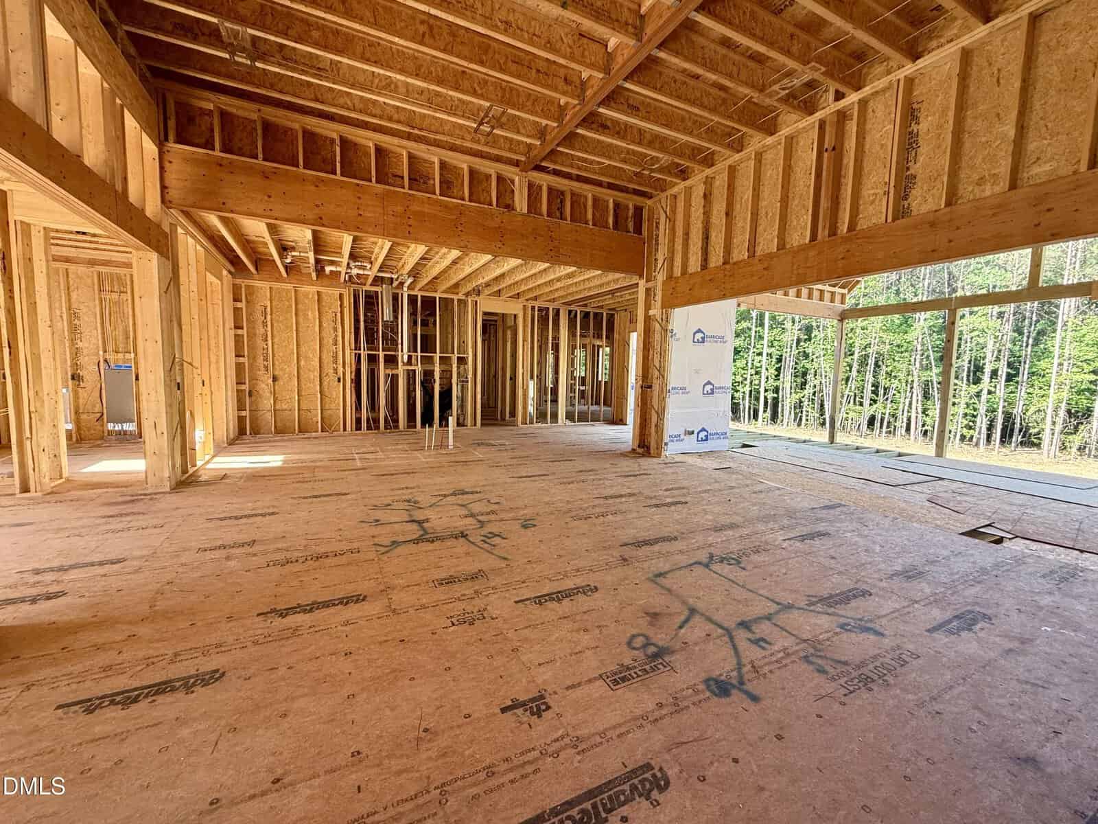Interior view of a house under construction, showing exposed wooden framing, open floor plan, and sunlight streaming through large windows. Blue markings are visible on the plywood floor. Trees can be seen outside.