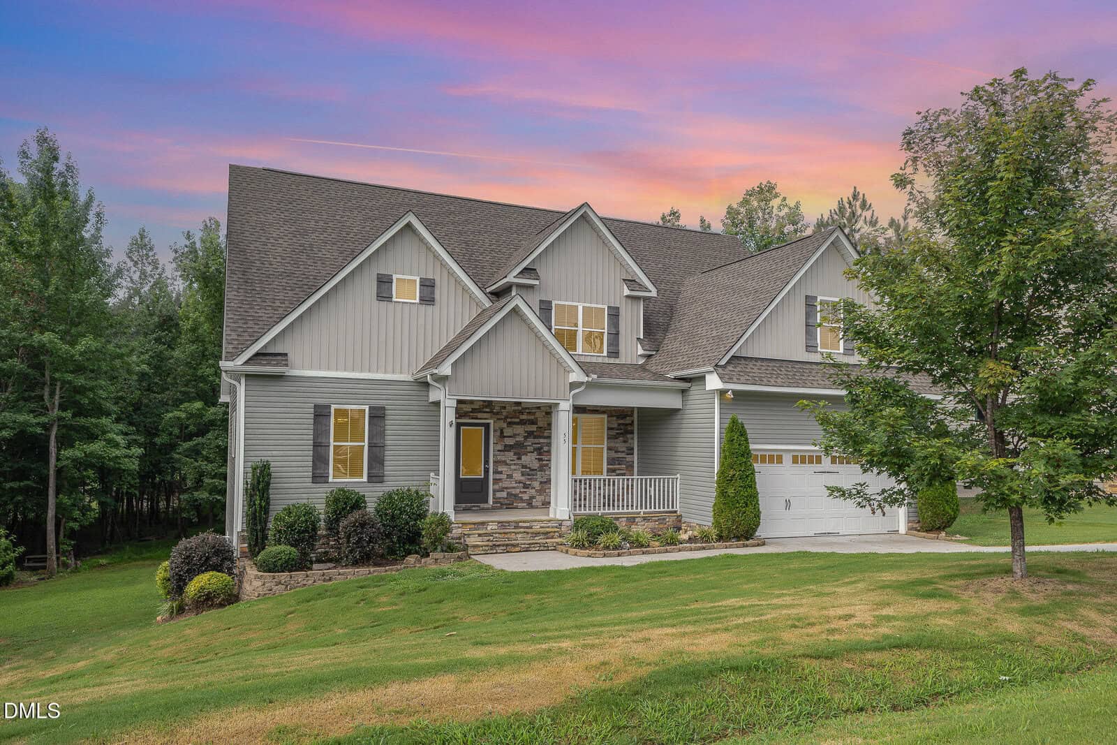 Gray two-story house with front porch, gabled roof, and attached white garage, surrounded by green lawn and trees, with a vibrant pink and purple sunset sky in the background.