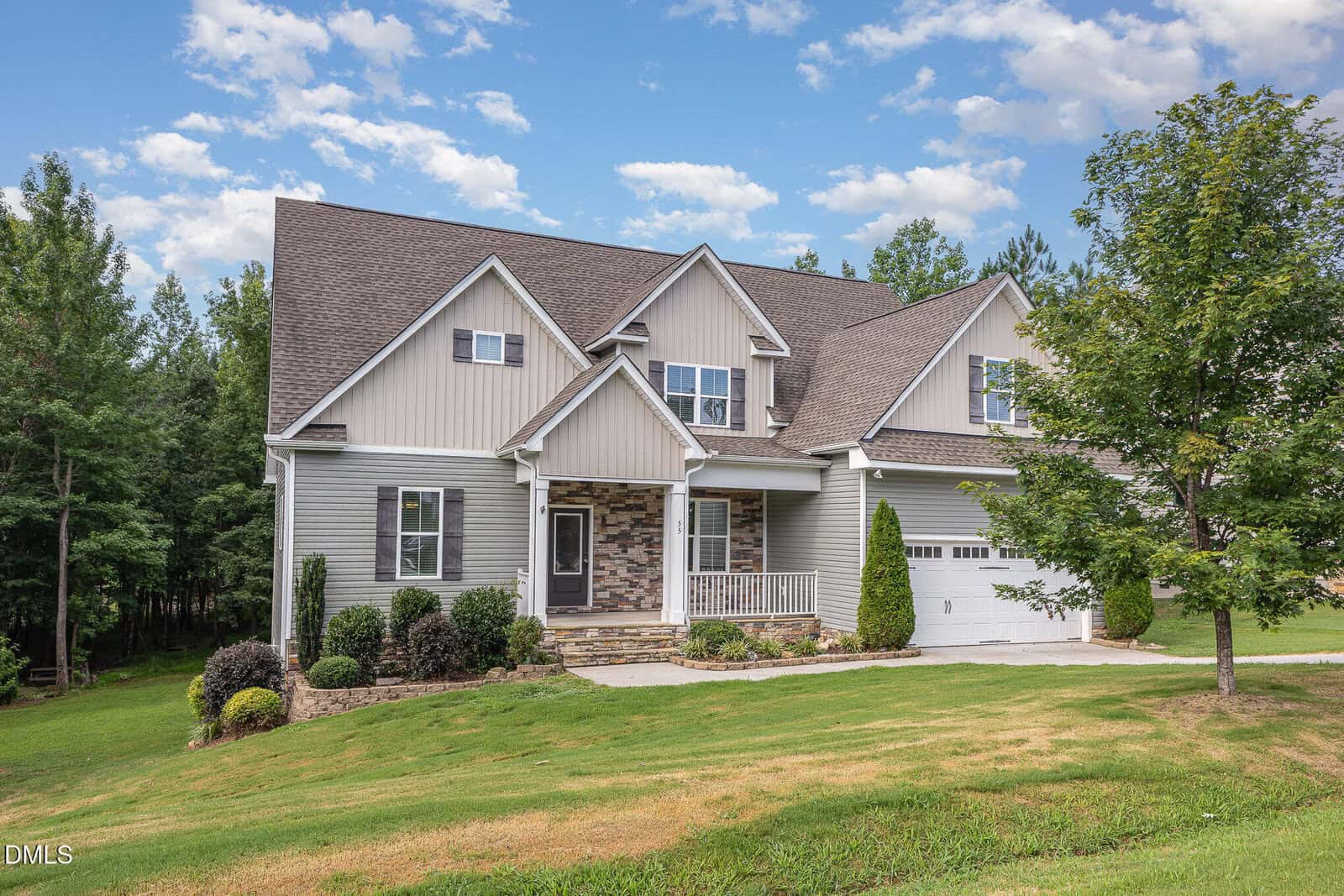 A two-story house with tan siding, stone accents, a covered front porch, attached garage, landscaped yard, and a tree in front, set against a backdrop of trees and a partly cloudy sky.