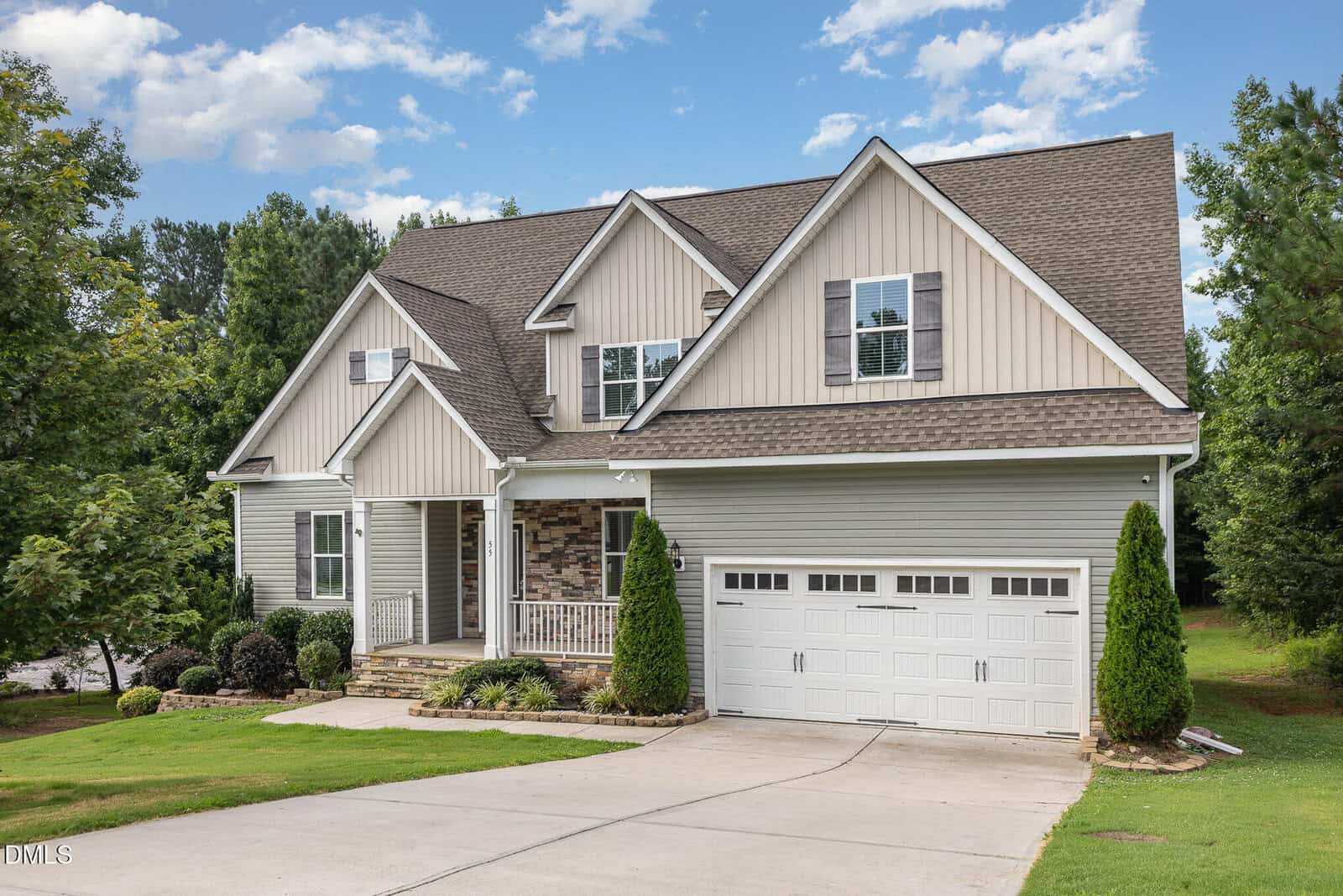 Two-story suburban house with beige siding, stone accents, white trim, and a double garage. There are small shrubs and landscaping in front, with a green lawn and trees in the background under a partly cloudy sky.