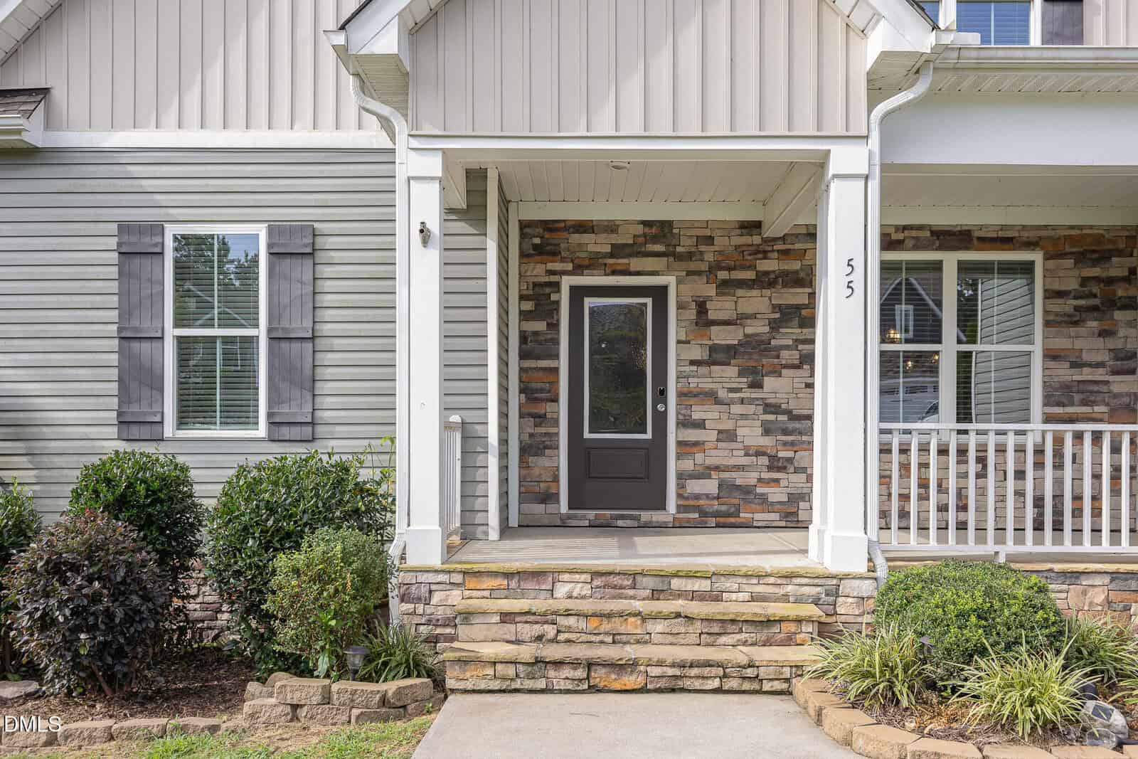 The image shows a house entrance with stone steps leading to a covered porch, a dark front door with glass panel, gray siding, stone accents, and trimmed bushes along the walkway. The house number 55 is visible near the door.