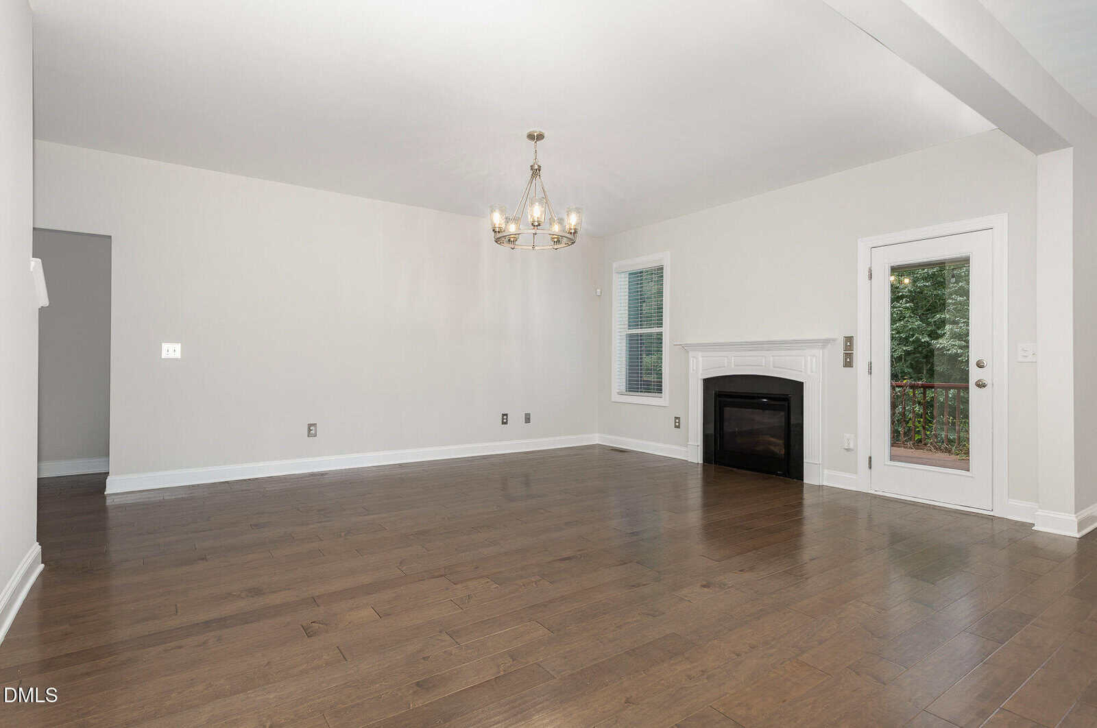 A spacious, empty living room with hardwood floors, neutral walls, a modern chandelier, a fireplace with a white mantel, large windows, and a glass door leading to a deck with greenery outside.