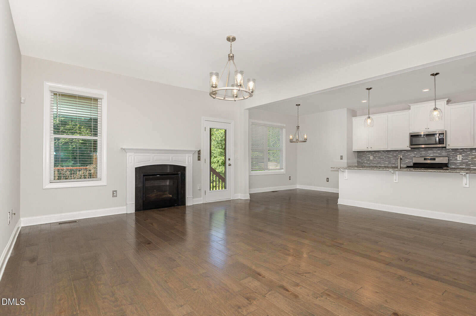 Bright, empty living area with hardwood floors, a white fireplace, and large windows. The open floor plan connects to a modern kitchen with white cabinets, stainless steel appliances, and pendant lights.
