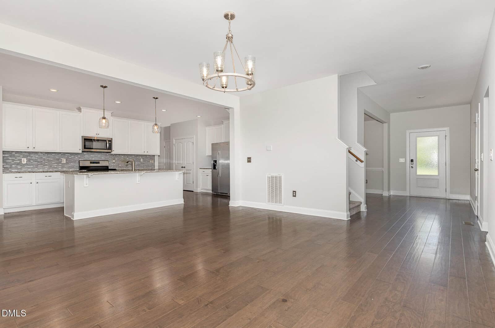 Modern open-concept kitchen and living area with hardwood floors, white cabinets, stainless steel appliances, pendant lights over an island, and a chandelier. A staircase and hallway lead to a door in the background.