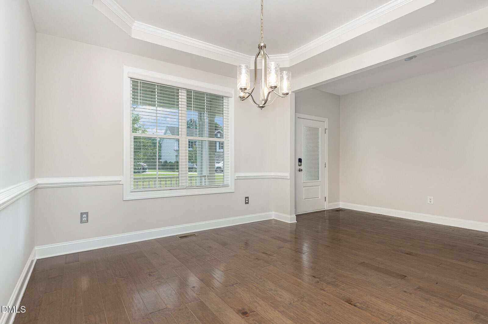 Bright, empty room with light gray walls, white trim, hardwood floors, a window with blinds, a modern chandelier, and a white front door. Natural light enters through the window, illuminating the space.