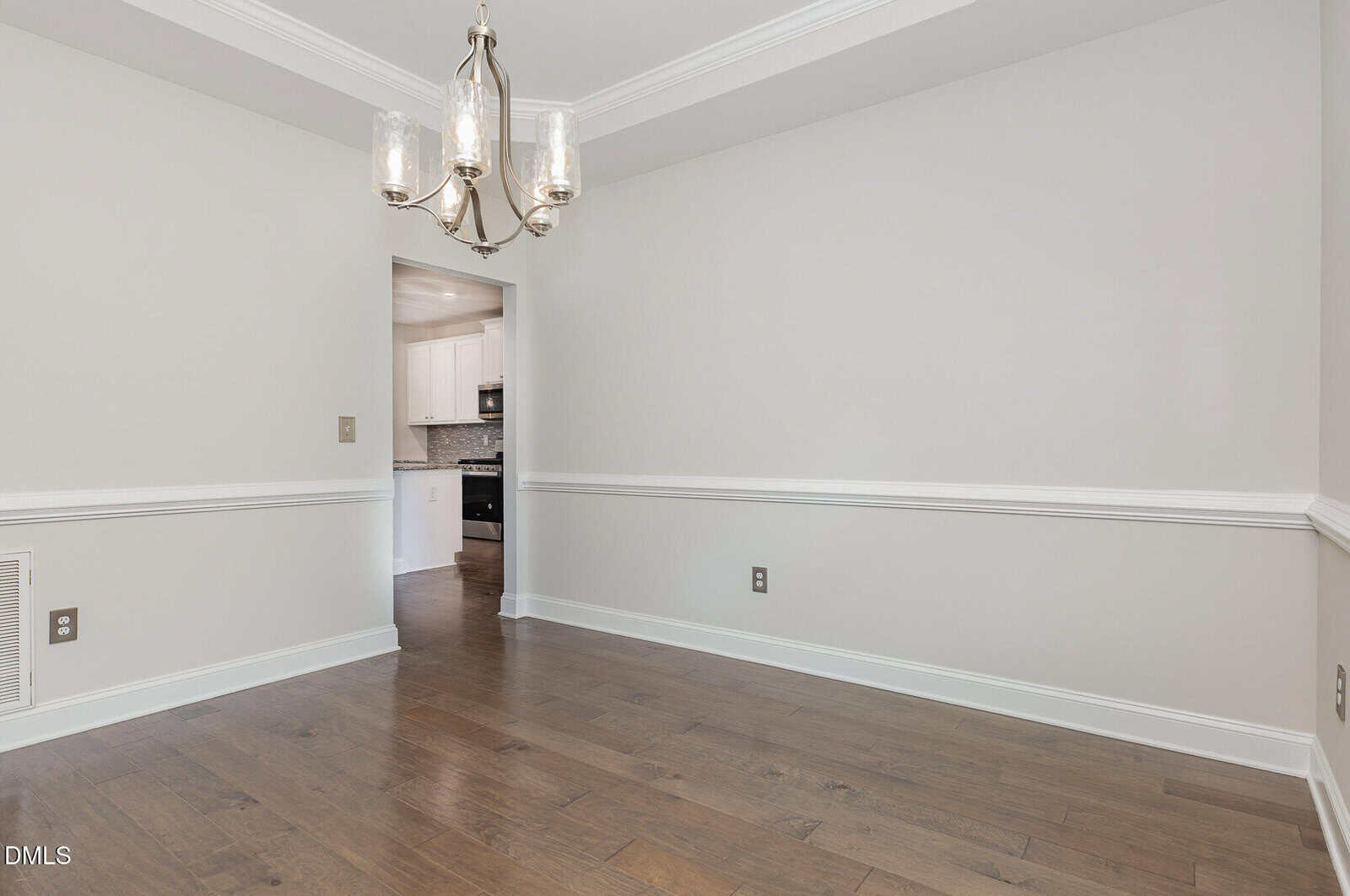A bright, empty dining room with light gray walls, white trim, crown molding, hardwood floors, and a modern three-light chandelier. An open doorway leads to a kitchen with white cabinets and tile backsplash.