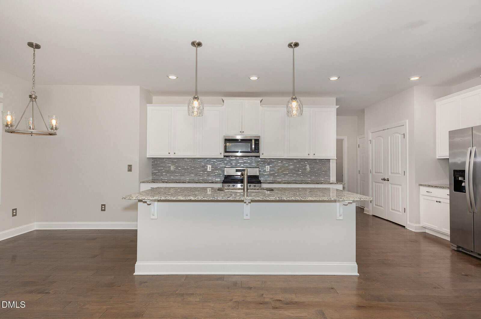 Modern kitchen with white cabinets, stainless steel appliances, granite countertops, a large island with pendant lights, mosaic tile backsplash, and dark wood floors. A chandelier hangs in the adjacent dining area.