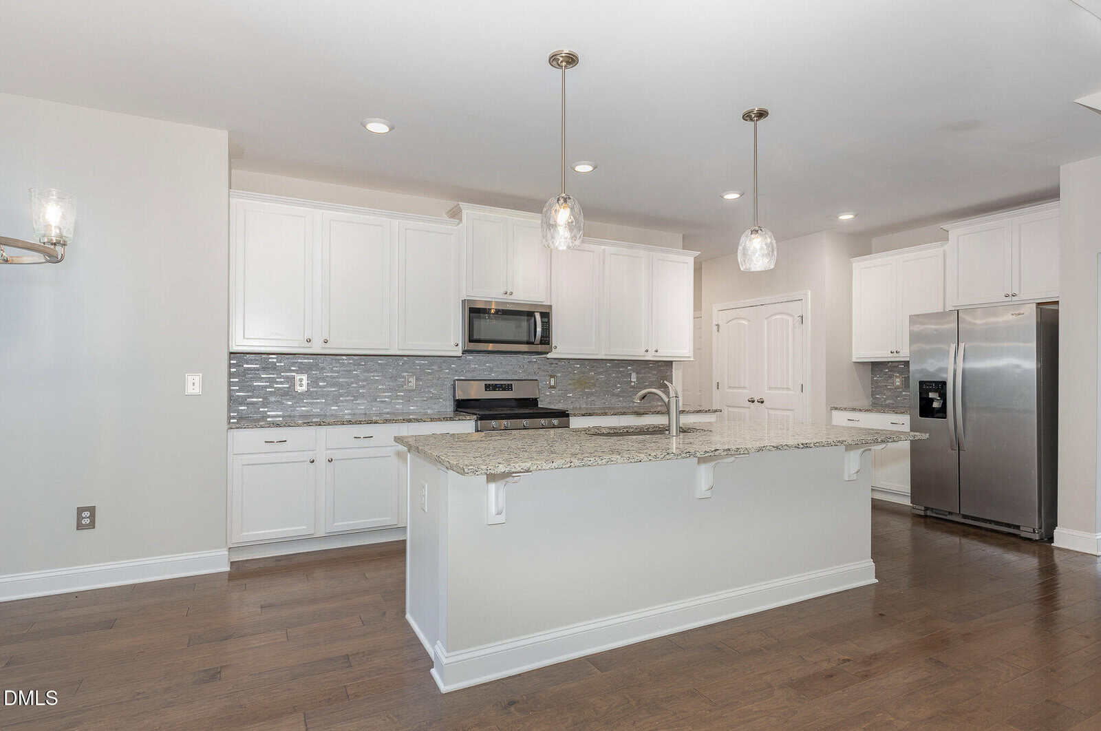 Modern kitchen with white cabinets, granite countertops, stainless steel appliances, a large island with sink, tile backsplash, pendant lights, and wood flooring.