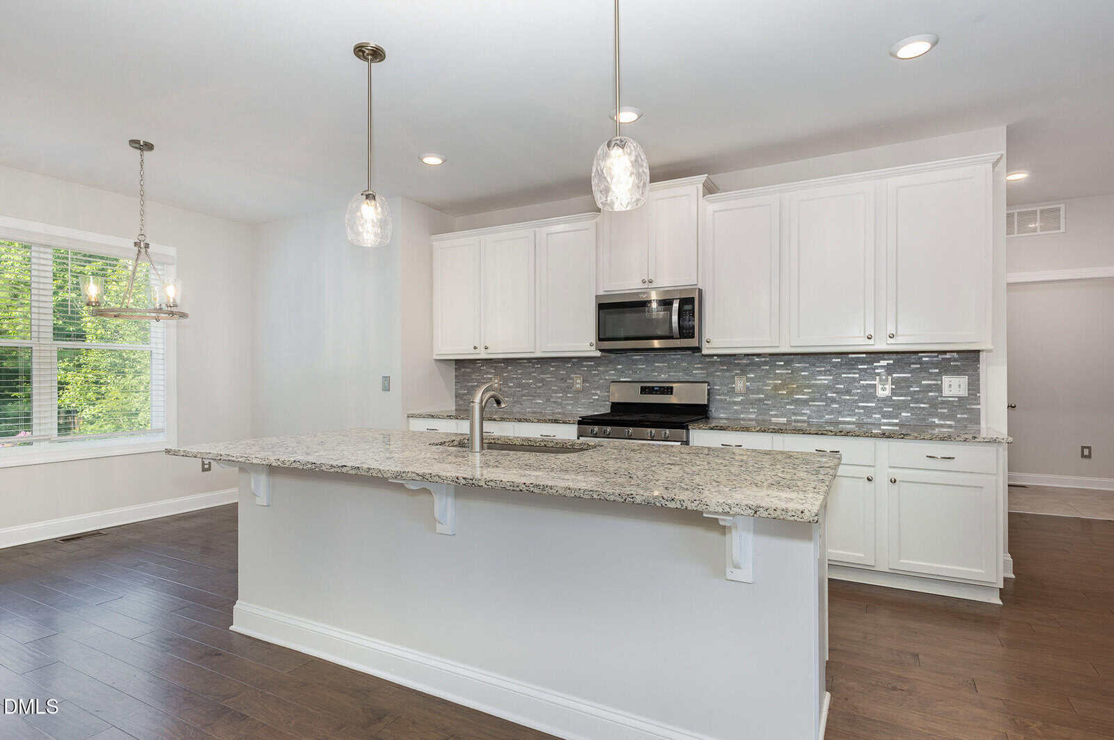 Modern kitchen with white cabinets, stainless steel appliances, a tile backsplash, pendant lighting, and a large granite island with a built-in sink. Hardwood floors and a dining area with large windows are visible.