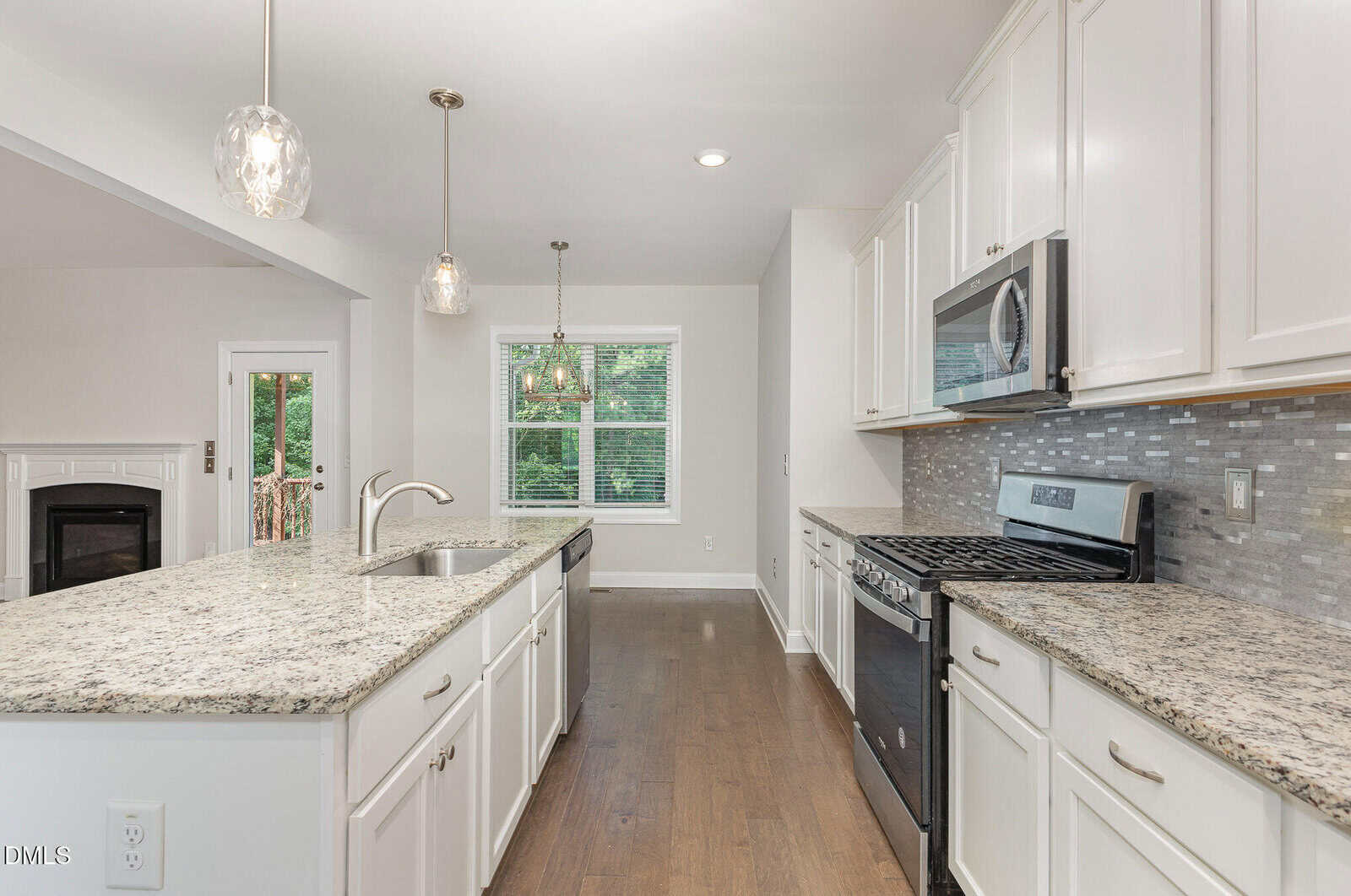 Modern kitchen with granite countertops, white cabinets, stainless steel appliances, pendant lights over an island, tile backsplash, and a window overlooking greenery. Wooden floors and a fireplace are also visible.