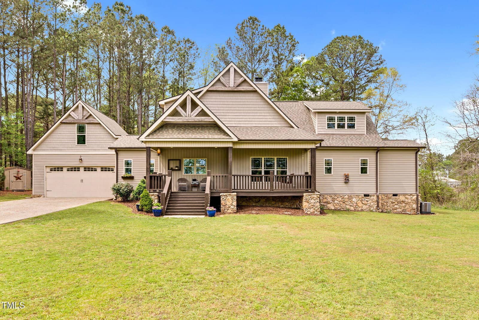 A large modern farmhouse with beige siding, stone accents, a covered front porch, and an attached two-car garage, set on a spacious, grassy lawn bordered by tall pine trees.