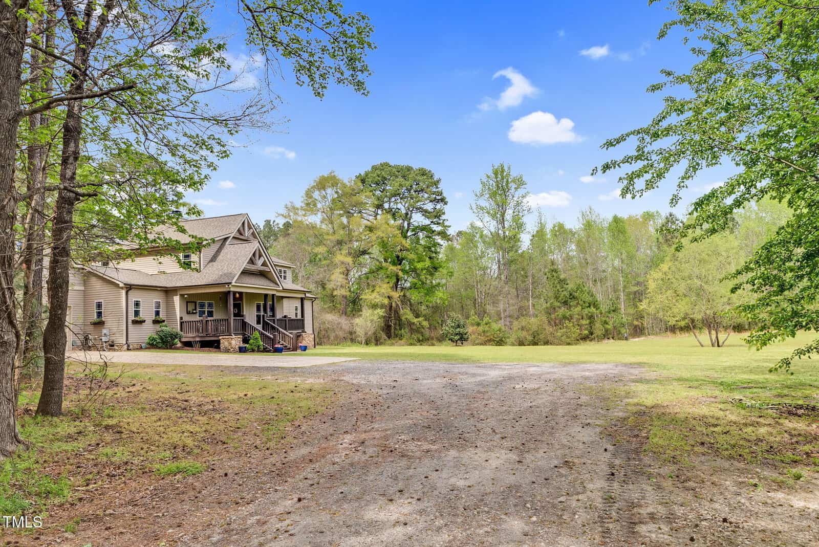 A beige house with a covered porch sits beside a gravel driveway, surrounded by trees and greenery under a clear blue sky.
