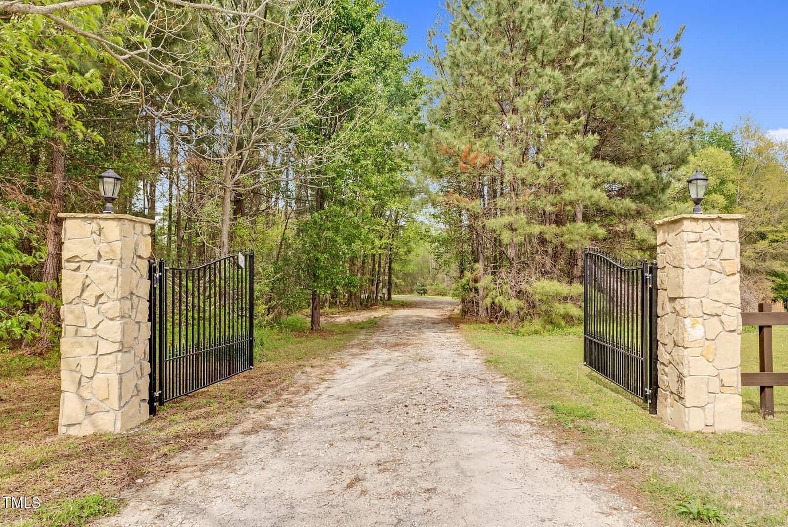 A gravel driveway passes between two stone pillars with black iron gates, leading into a wooded area with green trees and pine trees on both sides under a clear blue sky.