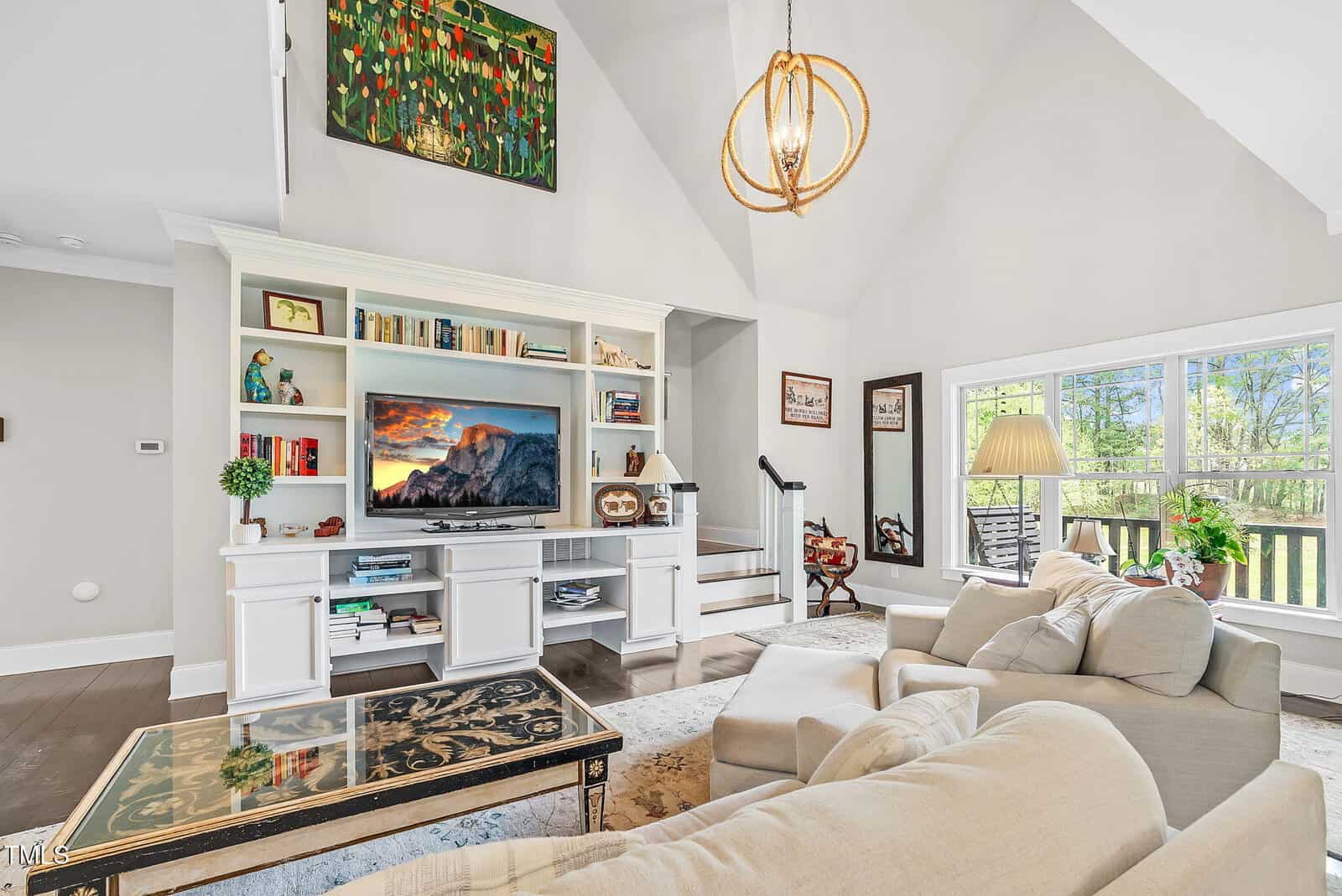 Bright living room with high ceilings, a white entertainment center with a TV, shelves, and decor, beige couches, a patterned coffee table, large windows, and modern light fixture. Stairs and artwork are visible in the background.