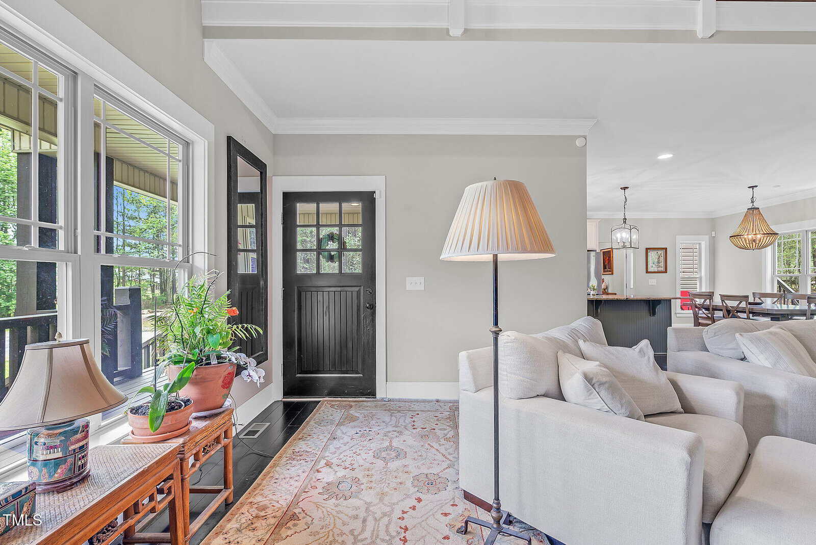 A bright living room with light gray walls, a patterned area rug, beige sofas, floor and table lamps, a black front door, large windows, potted plants, and a view into a dining area with a chandelier.
