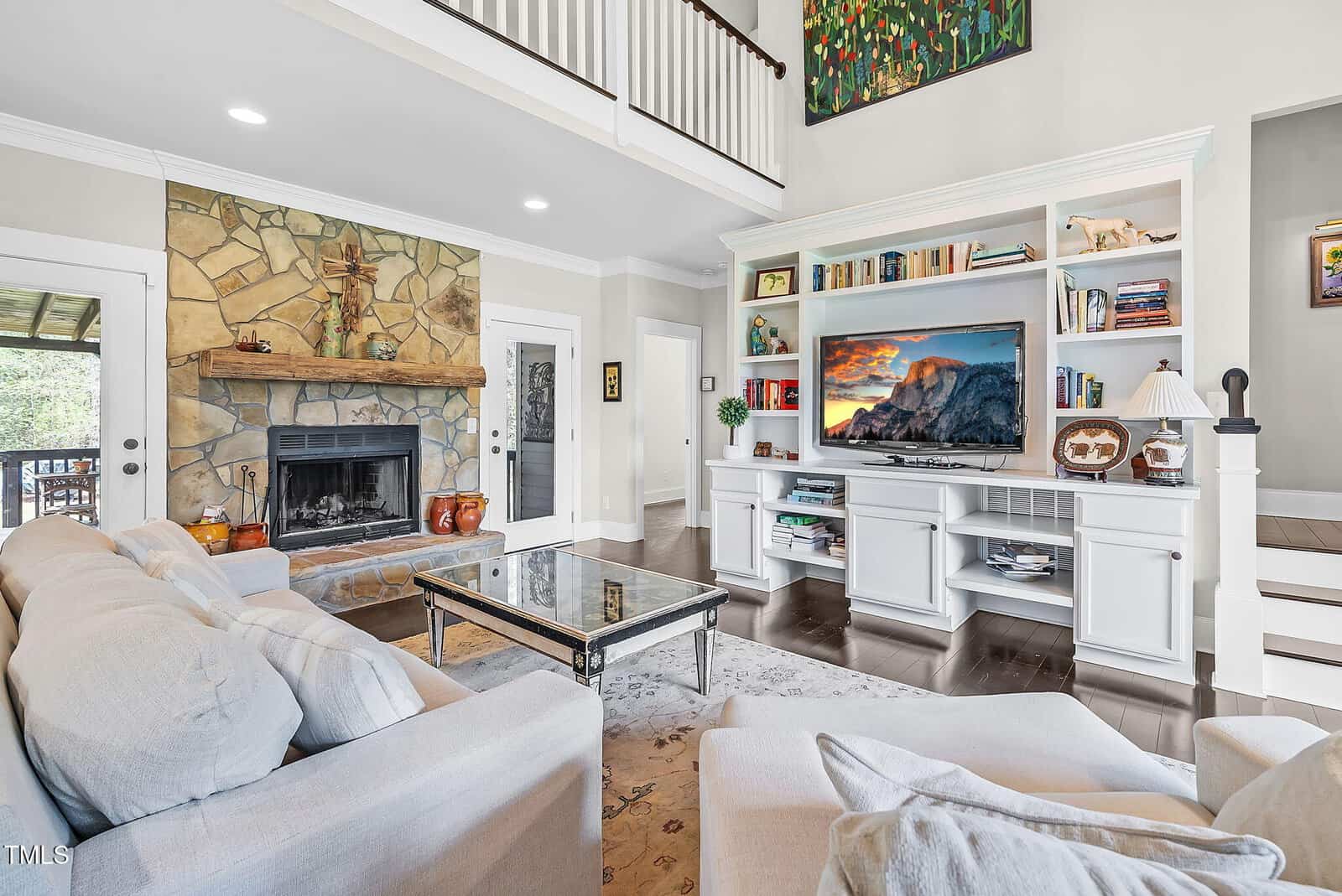 A bright, open living room with a stone fireplace, white built-in shelves, a TV, cozy light gray sofas, a glass coffee table, and a decorative rug. Stairs and a covered porch are visible in the background.