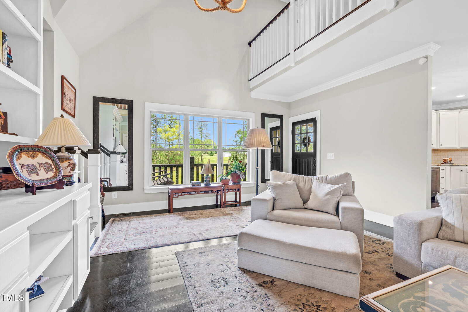 Bright living room with vaulted ceiling, large window letting in natural light, light-colored armchairs, built-in shelves with decor, a rug, and a view into the kitchen; greenery visible outside.
