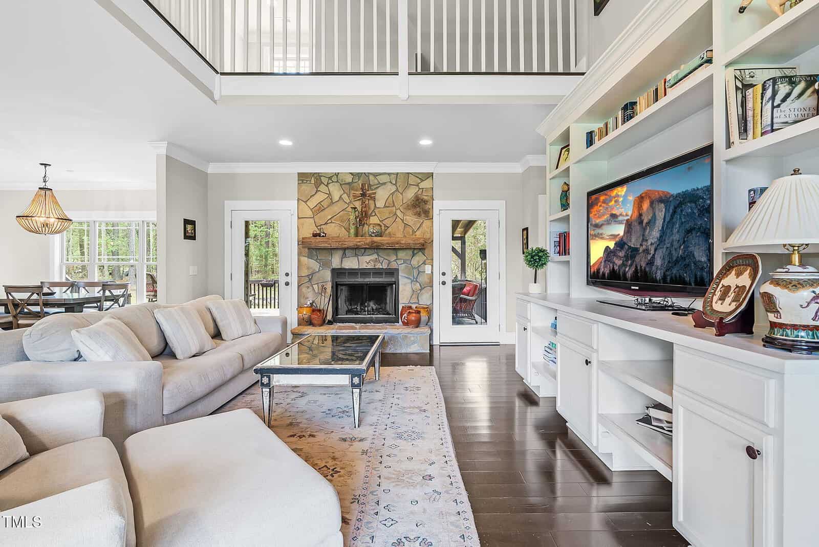 A bright, spacious living room with a sectional sofa, a glass coffee table, a stone fireplace, and built-in white shelves with a TV and books, connected to a dining area, featuring large windows and dark wood floors.