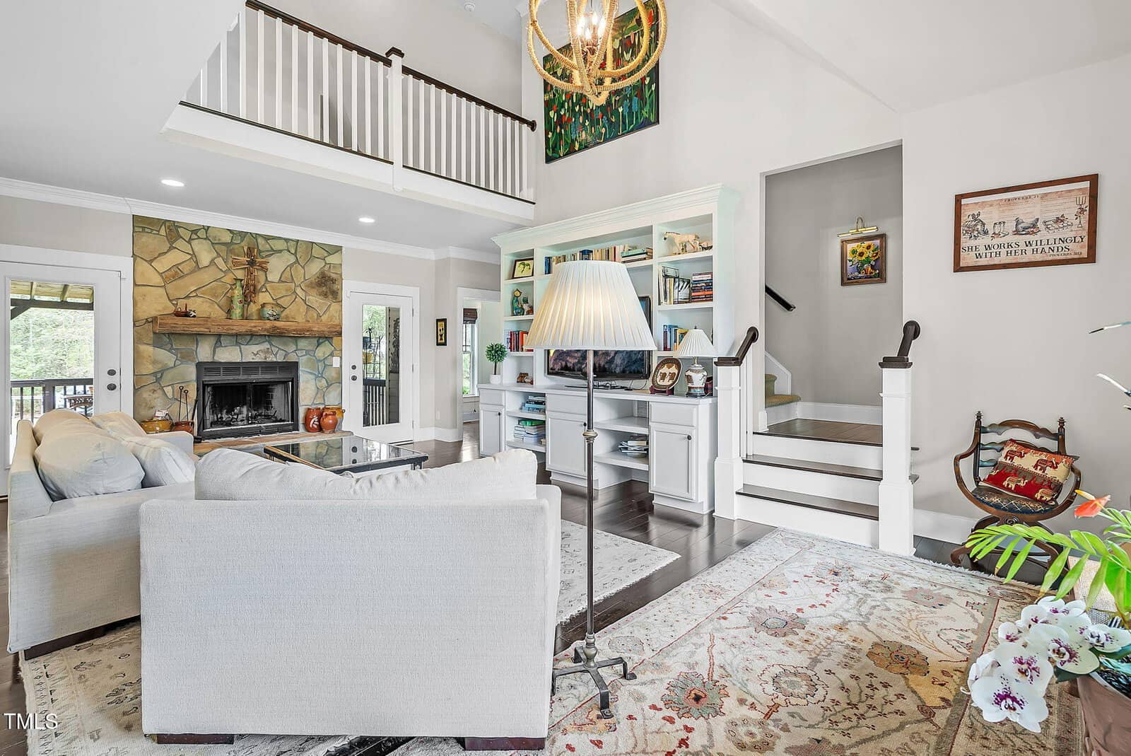 Bright living room with vaulted ceiling, white couch, stone fireplace, built-in shelves, staircase, indoor plants, a large rug, and natural light from glass doors and windows. Cozy, inviting, and stylish space.