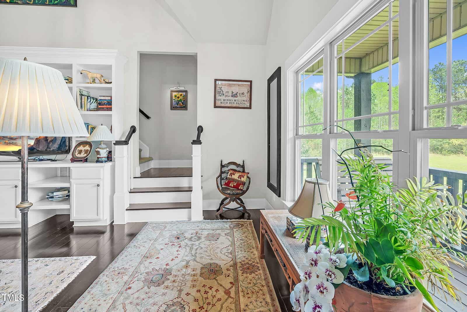 Bright living room with white walls, large windows, a floral rug, houseplants, a wooden table, built-in bookshelves, and stairs leading up. Sunlight fills the space, highlighting cozy and inviting decor.