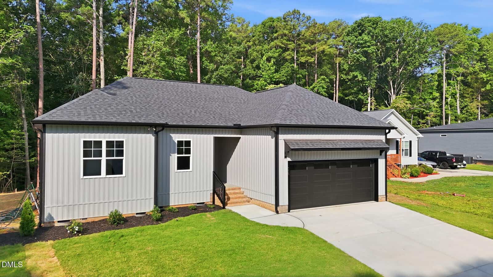 A modern single-story house with gray siding, black trim, and a black double garage door, set on a green lawn with a concrete driveway and surrounded by tall trees.