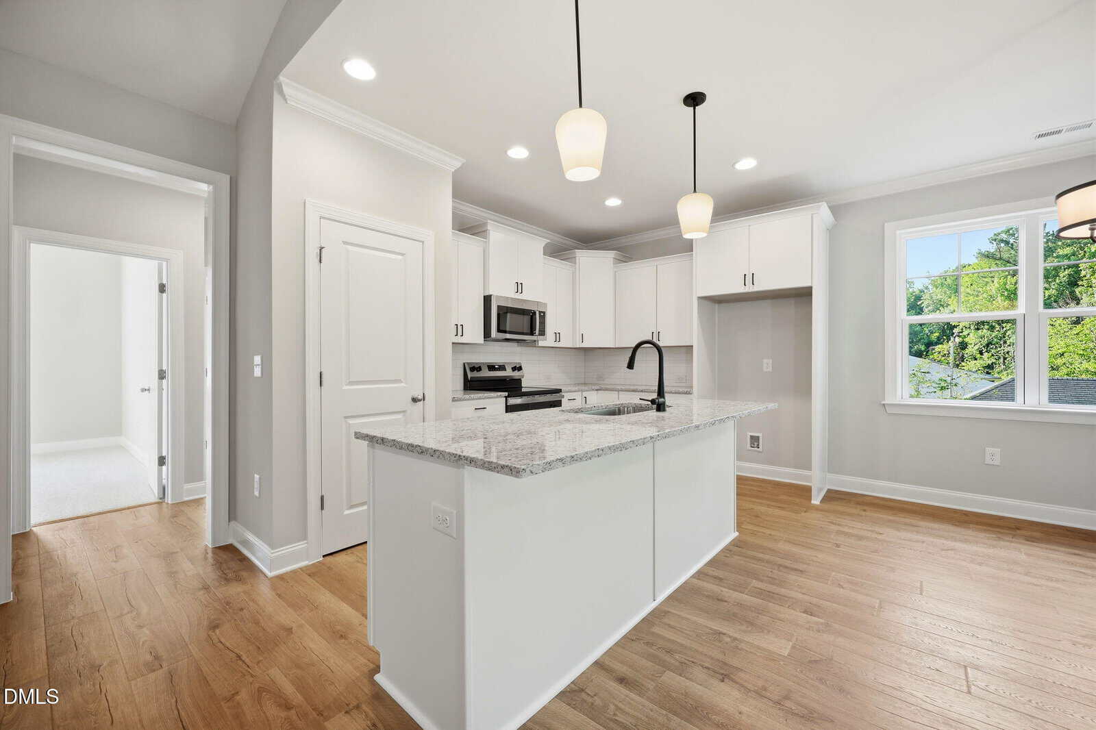 Modern kitchen with white cabinets, stainless steel appliances, a large island with a granite countertop and black faucet, light wood flooring, pendant lights, and natural light coming through a window.