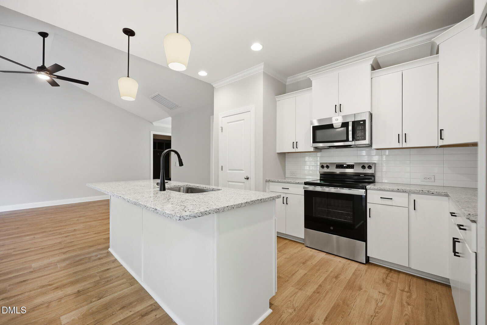 Modern kitchen with white cabinets, granite countertops, stainless steel appliances, an island with a sink, pendant lighting, and wood flooring. Adjacent open living space with ceiling fan visible.