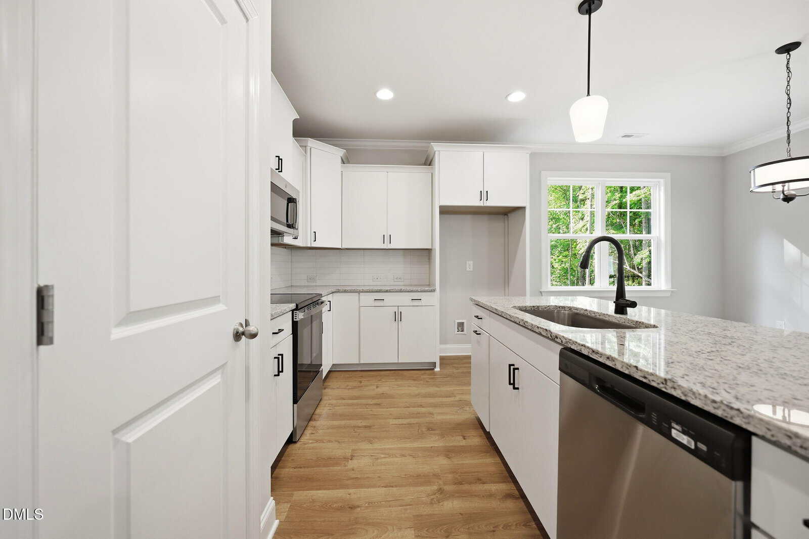 Modern kitchen with white cabinets, stainless steel appliances, a granite island with a black faucet, light wood flooring, and a window letting in natural light. A pendant light and chandelier hang from the ceiling.