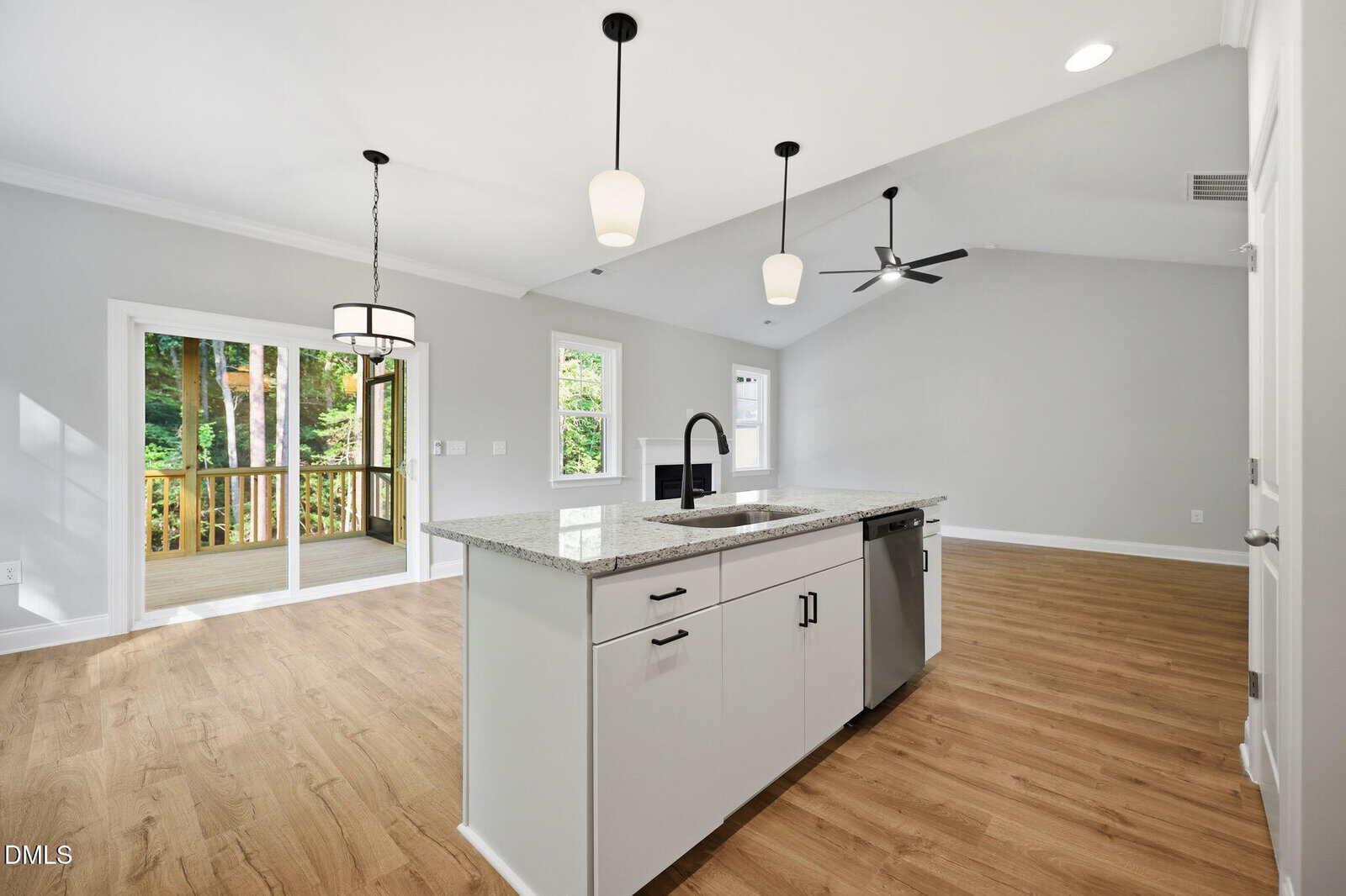 Modern open kitchen with white island, sink, and stainless steel dishwasher, overlooking a bright living area with wood floors, large windows, and sliding doors to a screened porch with views of trees outside.