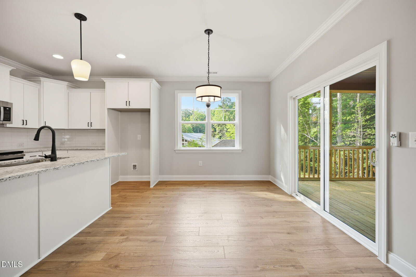 Bright kitchen and dining area with wood floors, white cabinets, granite countertops, pendant lights, large windows, and sliding glass doors leading to a covered wooden deck with a view of greenery outside.