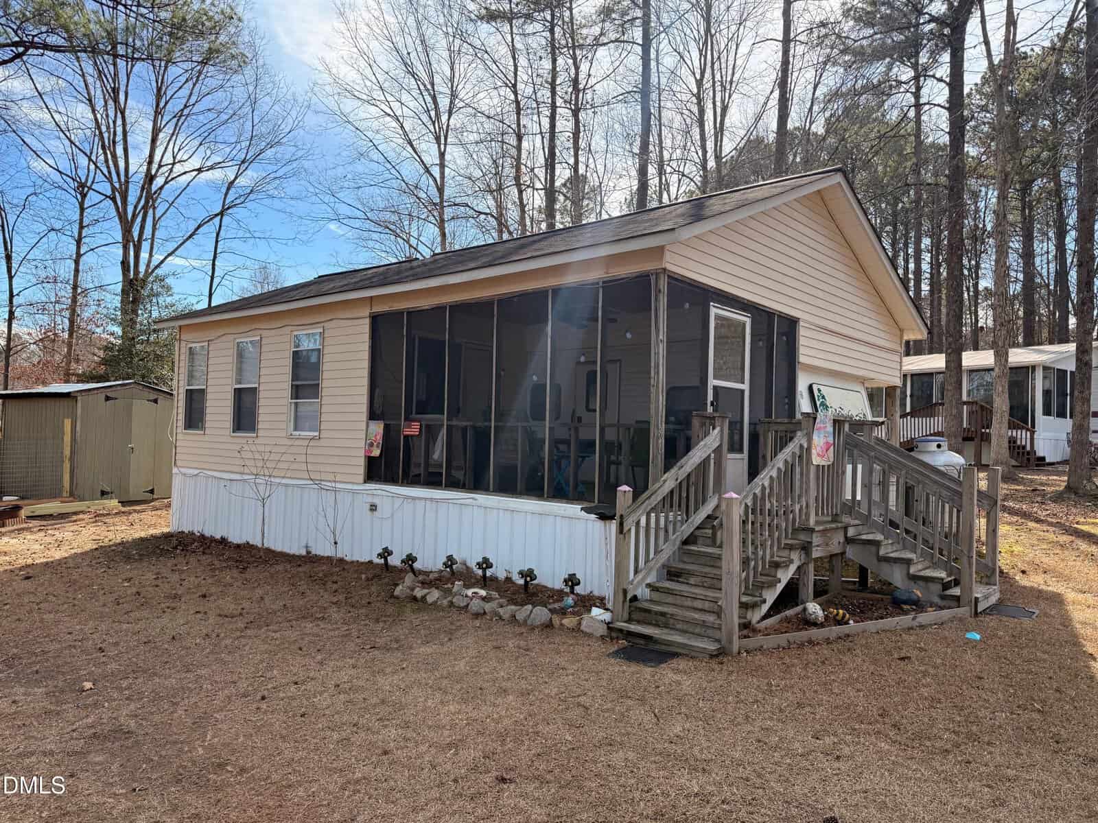 A tan mobile home with a screened-in porch and wooden stairs is situated in a wooded area at 136 Tomahawk Drive, Louisburg, with a small shed nearby and a lawn covered in dry grass.
