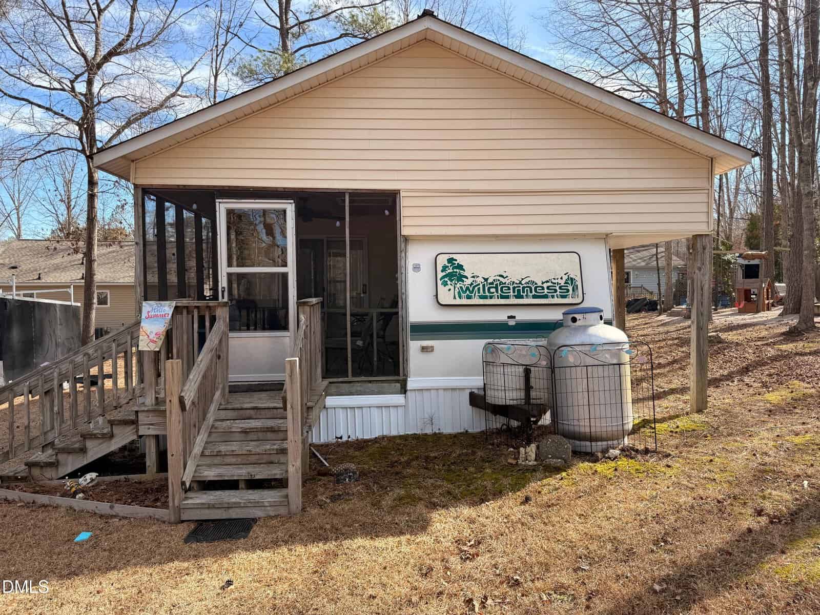 A small, single-story house with a screened porch, beige siding, wooden steps, and a “Wilderness” sign on the front. Two large propane tanks are visible beside the house, surrounded by leafless trees and dry grass.