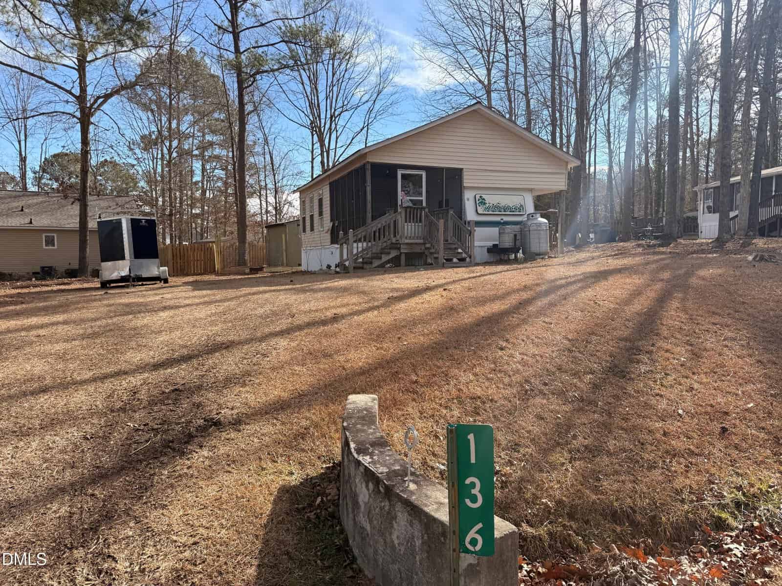 A small tan house with a front porch sits on a brown, grassy lot with tall trees. A green marker with the number 136 is at the front. There are neighboring houses and a trailer visible to the left.