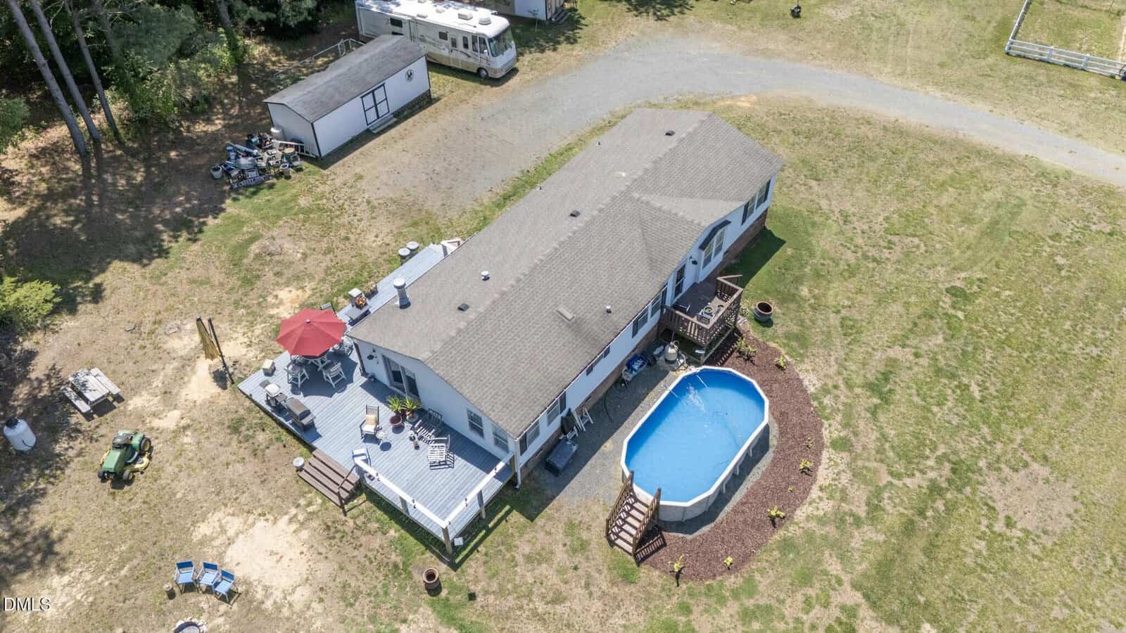 Aerial view of a single-story house with a large deck featuring a red umbrella, outdoor seating, and an above-ground oval pool in the backyard, surrounded by grassy land and trees.
