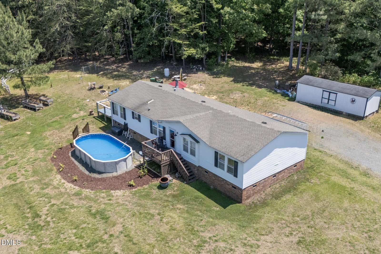 Aerial view of a single-story house with a gray roof, a small wooden deck, and an above-ground circular pool in the backyard, surrounded by grass, trees, and a white shed.