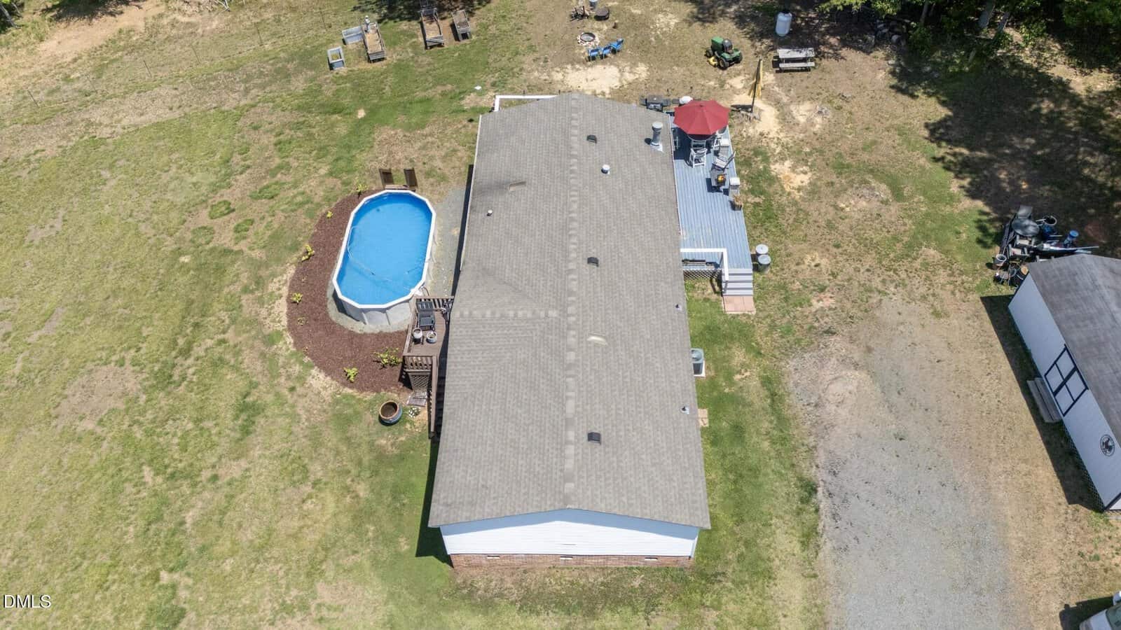 Aerial view of a house with a gray roof, raised deck with red umbrella, round above-ground pool, and surrounding yard with patches of grass and gravel area. Outdoor furniture and small structures are visible nearby.