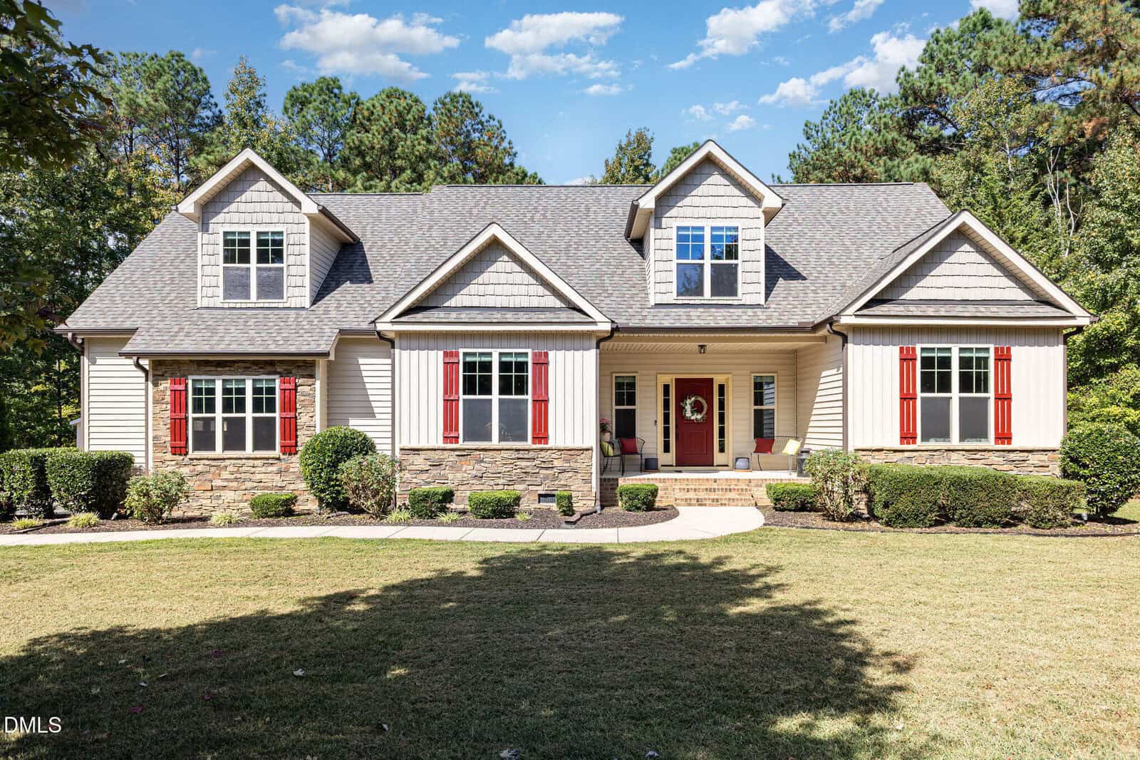A two-story suburban house with beige siding, stone accents, and red shutters, featuring a covered front porch, a manicured lawn, and surrounded by trees under a blue sky with scattered clouds.