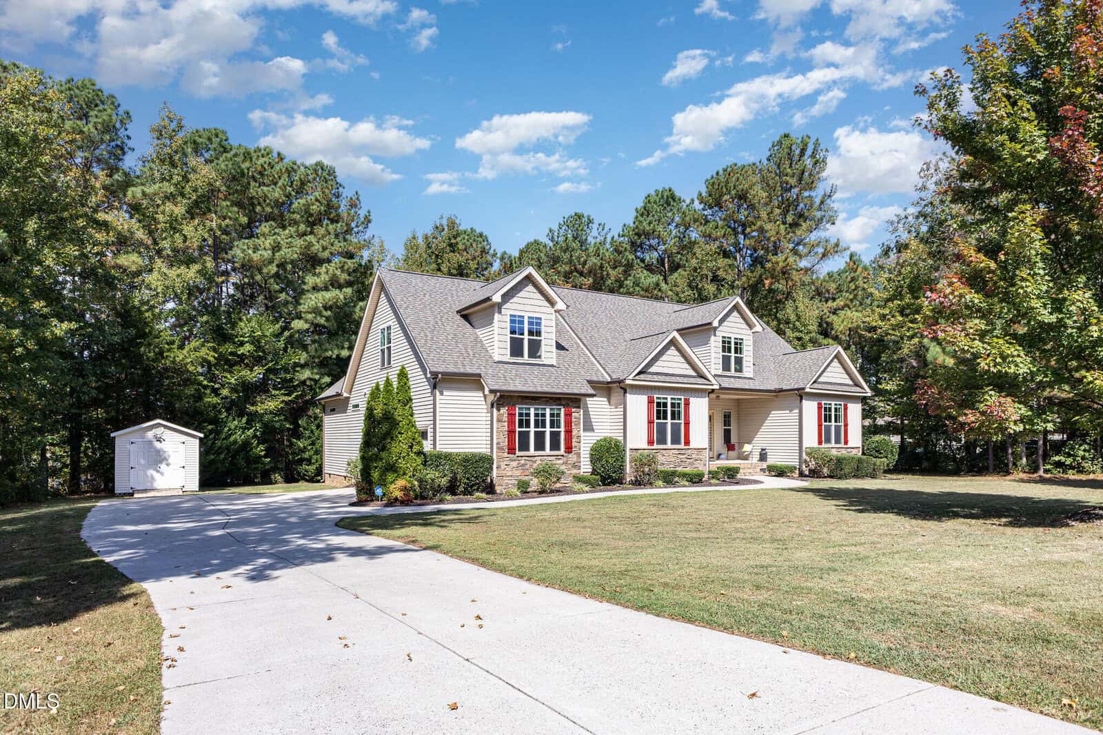 A two-story house with tan siding, red window shutters, and a gray roof sits on a spacious lot with a curved driveway, green lawn, small shed, and surrounded by trees under a partly cloudy sky.