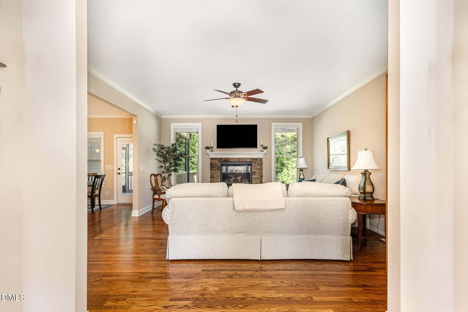 A living room with a light-colored sofa facing a fireplace and wall-mounted TV, hardwood floors, side tables with lamps, and large windows letting in natural light. A ceiling fan and an adjacent dining area are also visible.