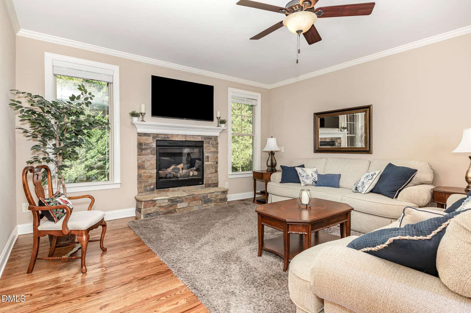 A living room with a beige sofa, armchair, wooden coffee table, and side tables. A stone fireplace with a black TV above it is centered between two windows. A ceiling fan and a potted plant are also visible.