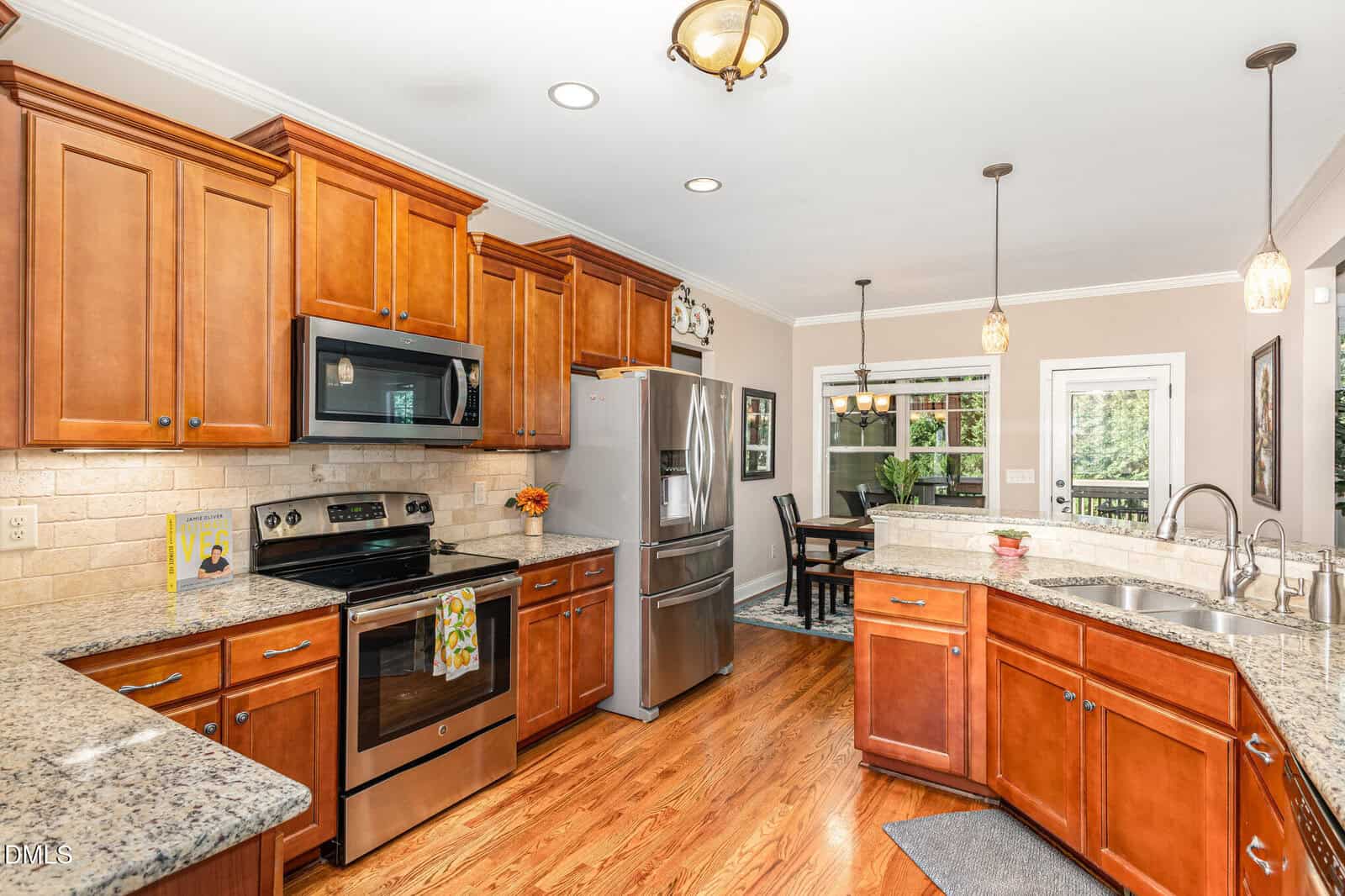 A modern kitchen with wooden cabinets, stainless steel appliances, granite countertops, a double sink, hardwood floors, and pendant lighting, adjacent to a dining area with a table and large windows.
