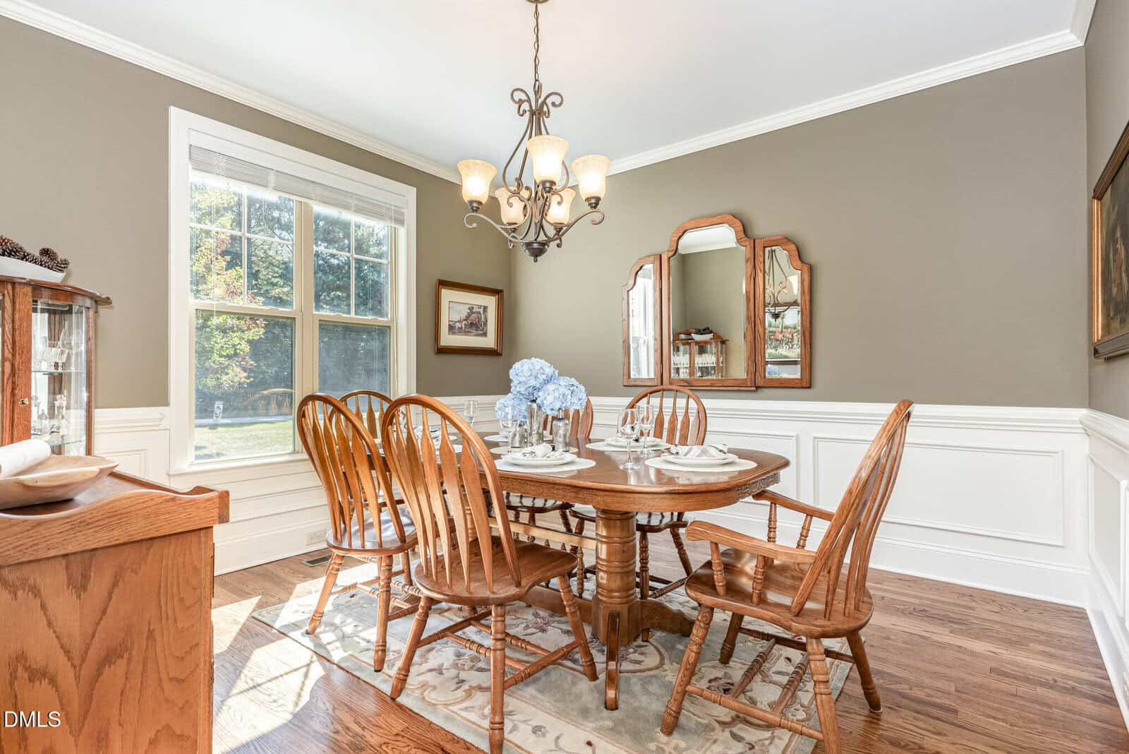 A dining room with a round wooden table set for six, wooden chairs, a chandelier, large window, and mirrors on the wall. Light streams in from the window, illuminating the hardwood floors and neutral-toned walls.