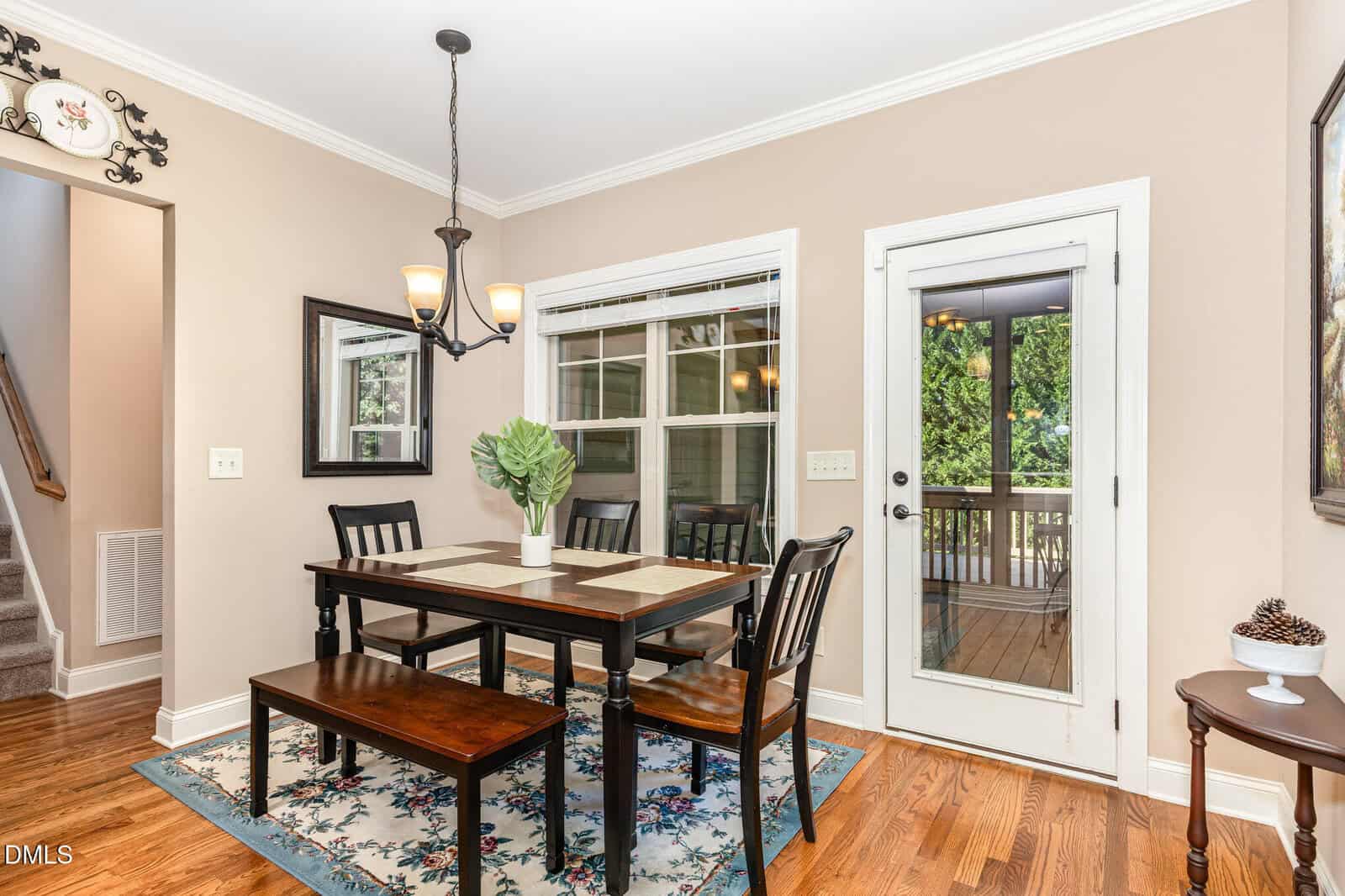 A dining room with a wooden table, four chairs, and a bench on a patterned rug. A chandelier hangs above the table. There is a glass door leading to a porch and a window overlooking greenery.